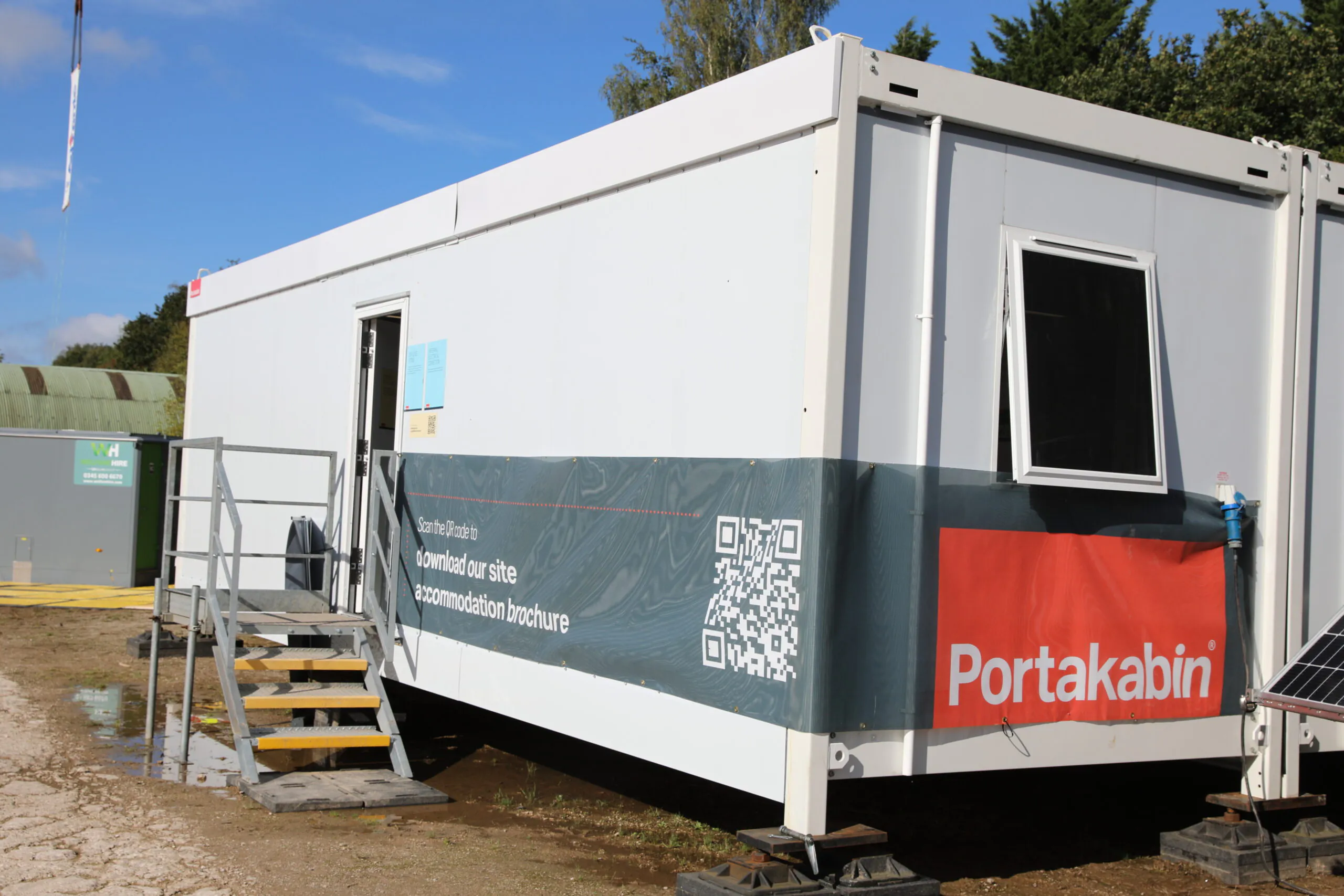 White portable building with steps leading to an entrance door, featuring a QR code and signage that reads "Download our site accommodation brochure." The brand "Portakabin" is displayed in red, indicating temporary site office or accommodation use.