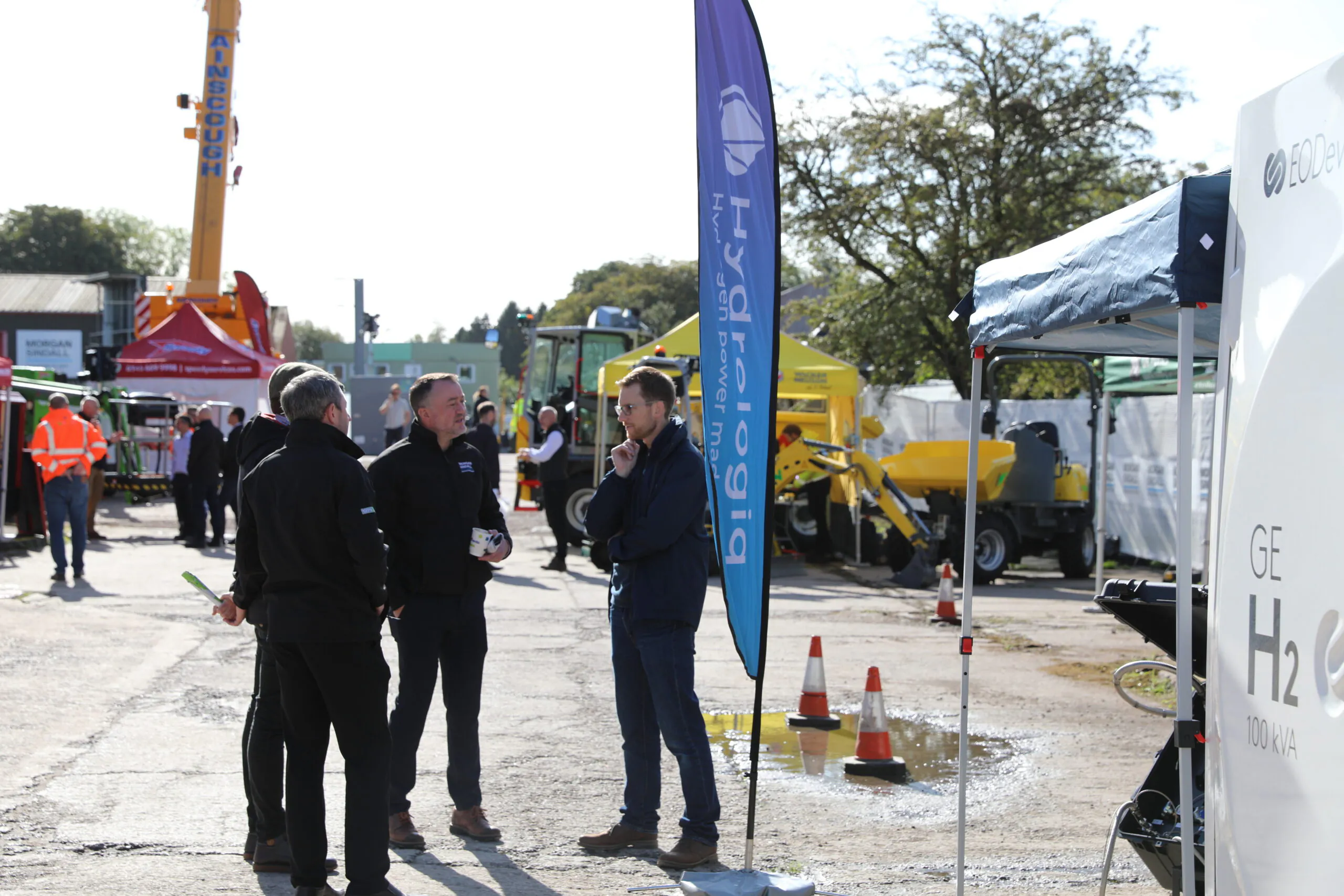 Several individuals conversing outdoors near industrial equipment setups; one person holds informational material.