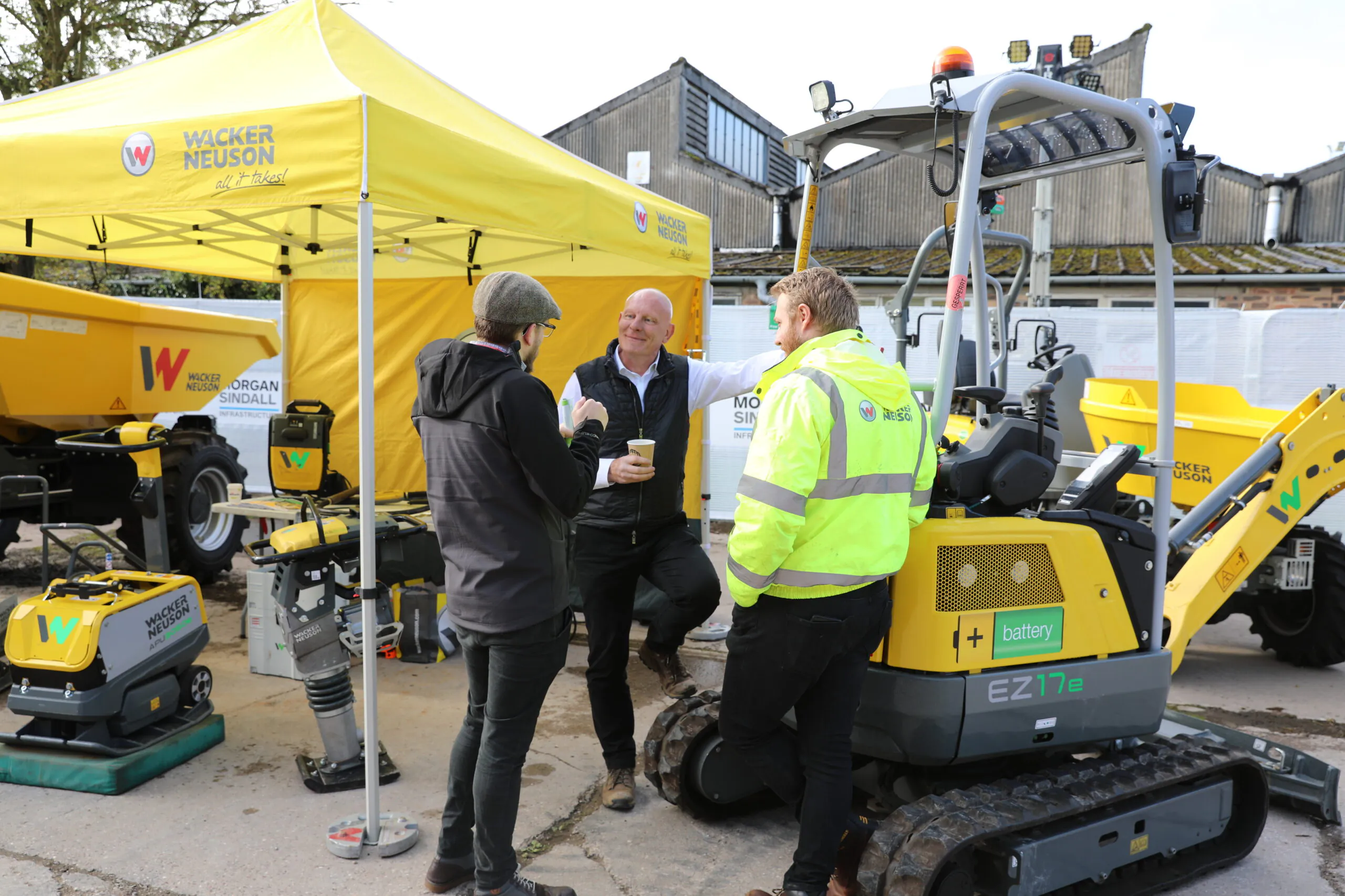Person in a bright yellow jacket standing under a yellow "Wacker Neuson" tent, appearing to be in conversation.