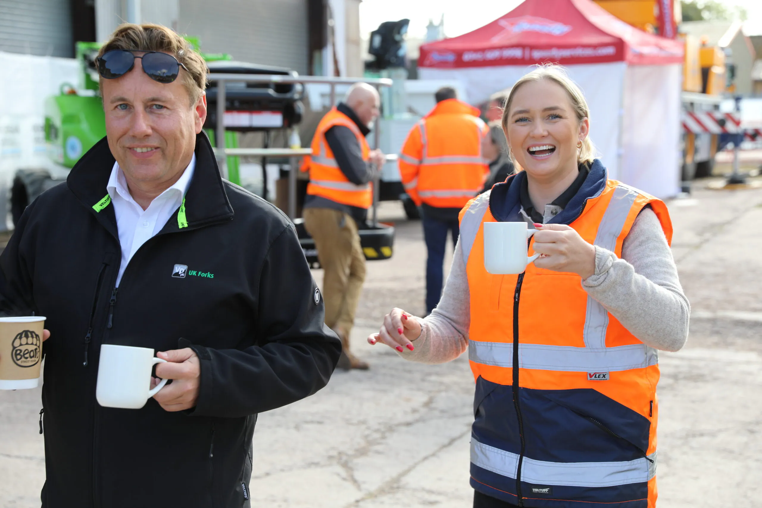 Two individuals in high-visibility vests smiling and holding mugs, with other workers in similar attire in the background.