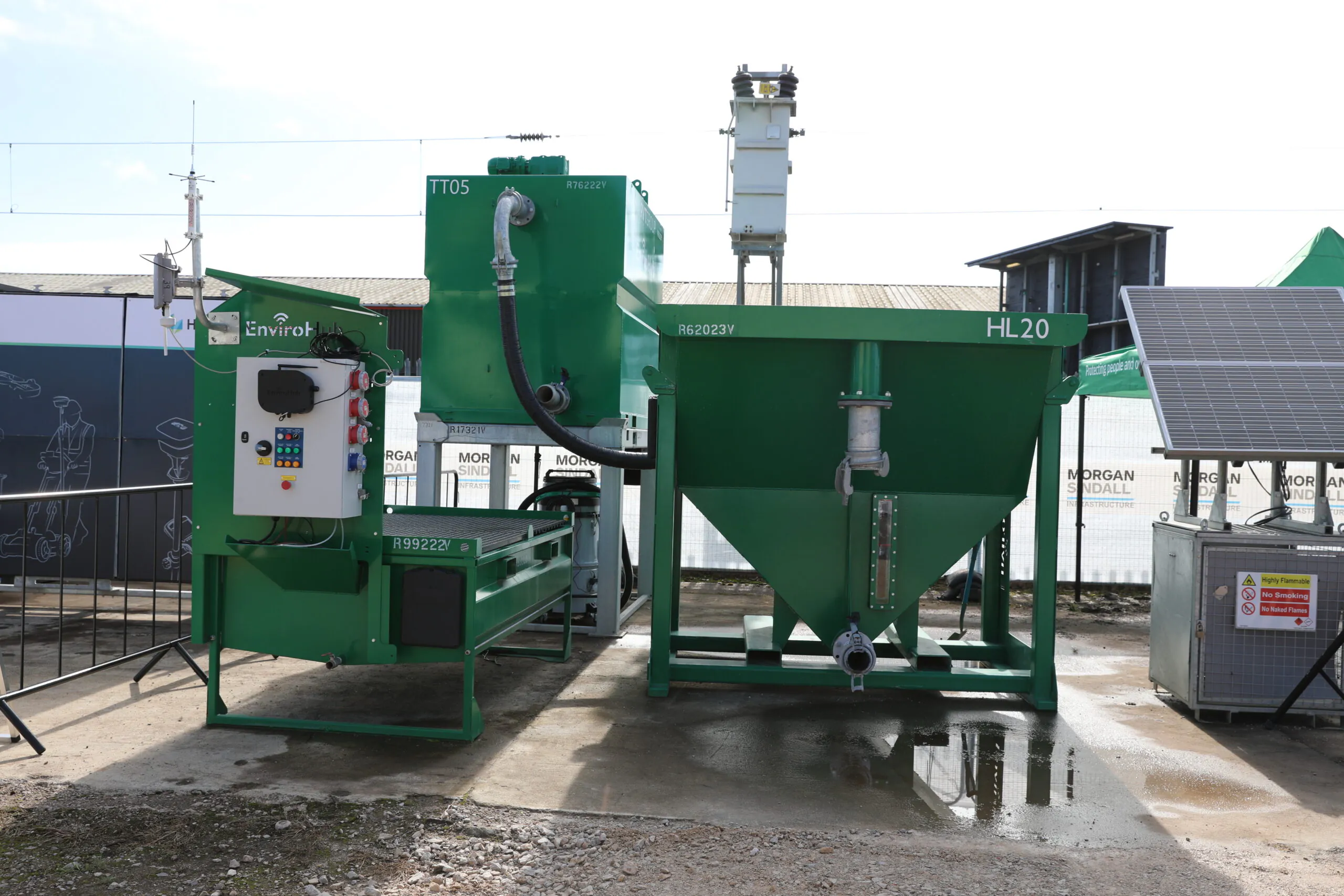 Outdoor setup of green industrial equipment labeled "IT05" and "HL20," part of an environmental or waste management system. The left unit features a control panel, and the right unit is a hopper with a pipe. Branding includes "EnviroH."