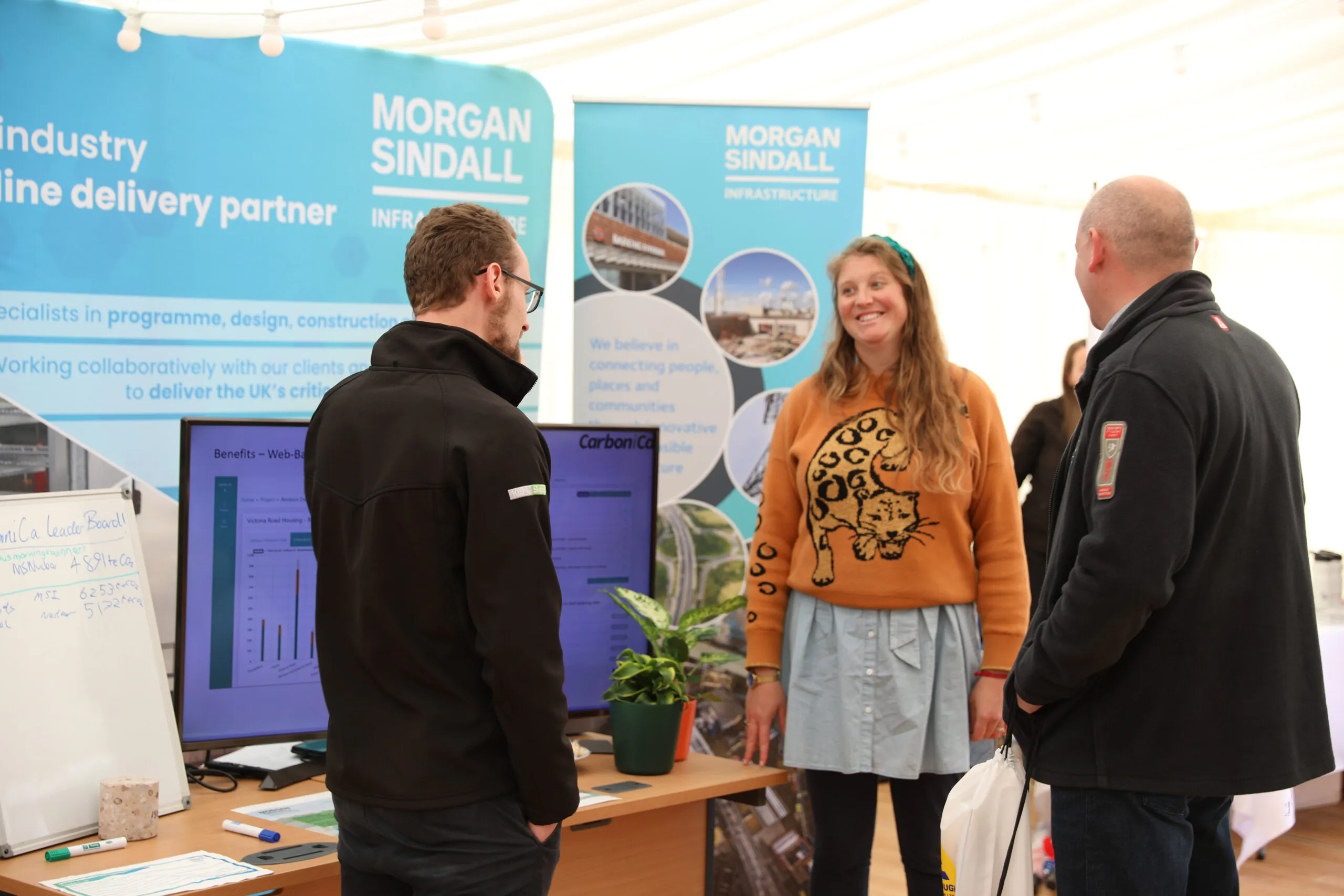 Three people conversing at a booth with "Morgan Sindall" banners, a TV screen displaying information, a small plant, and a whiteboard.