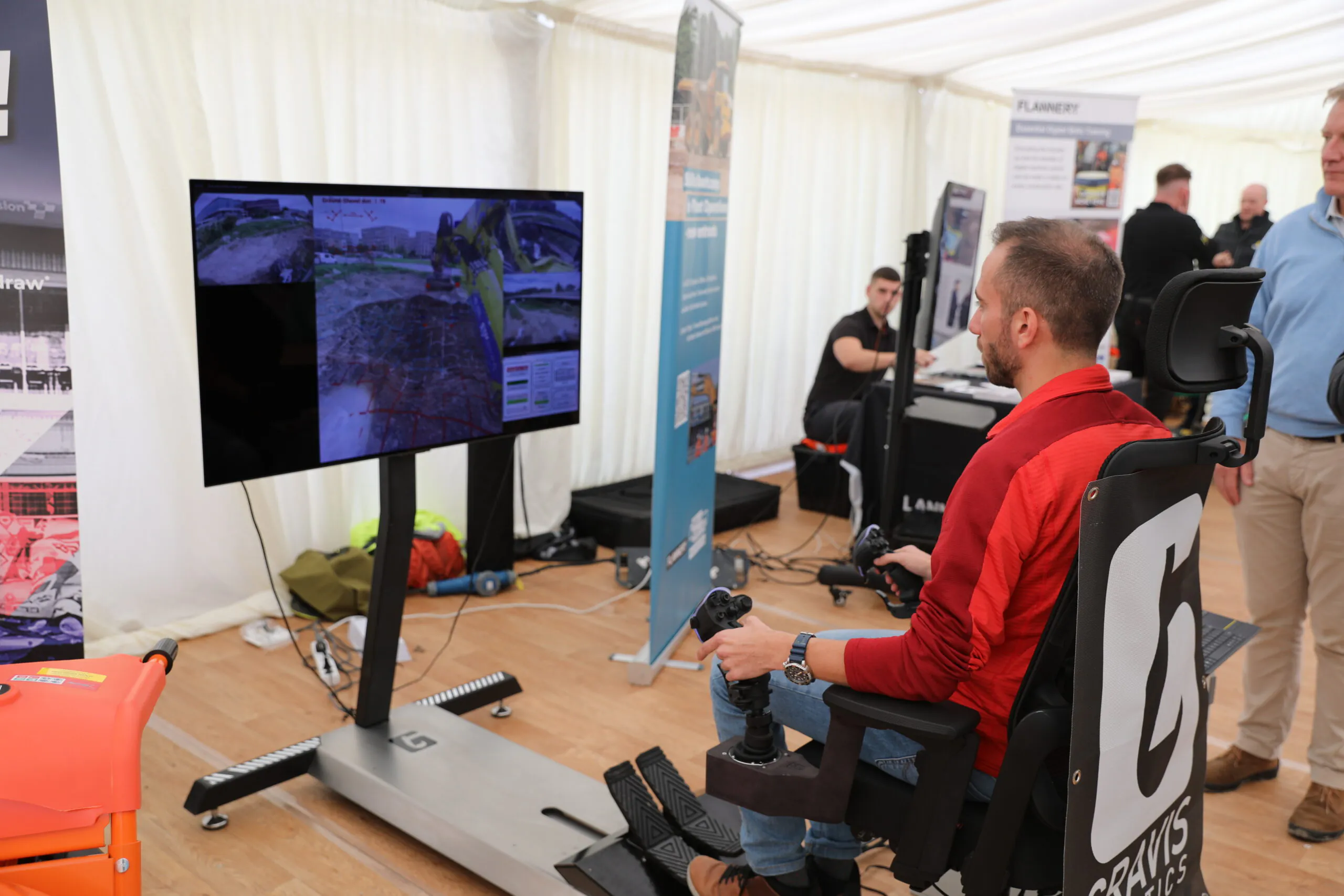 Individual seated in a simulator chair operating controls while viewing construction site visuals on multiple screens.