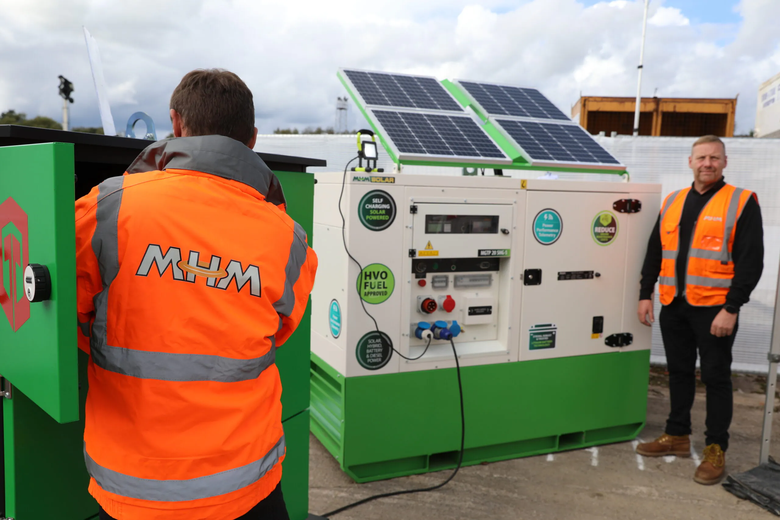 Two individuals in high-visibility jackets near a green and white machine with solar panels labeled for HVO fuel use. One person wears an orange "MHM" jacket. The equipment promotes low emissions and appears to be an eco-friendly power generator.