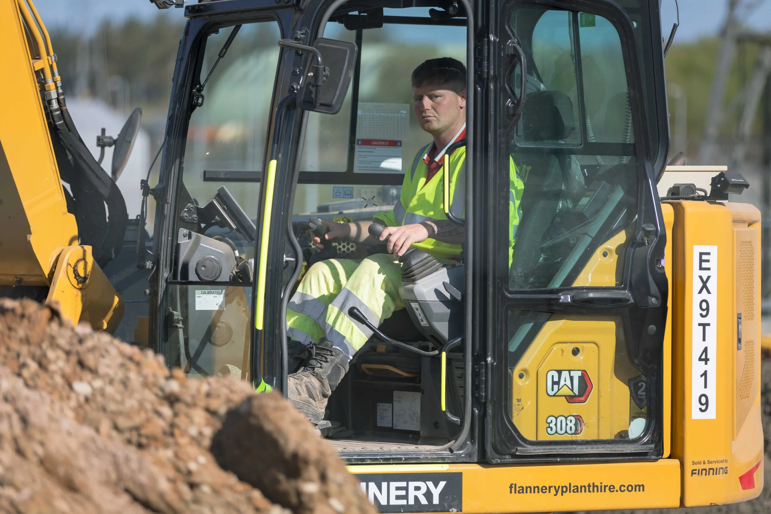 Construction worker operates a yellow CAT 308 excavator labeled "EX9T419," seated inside the cab and wearing high-visibility clothing.