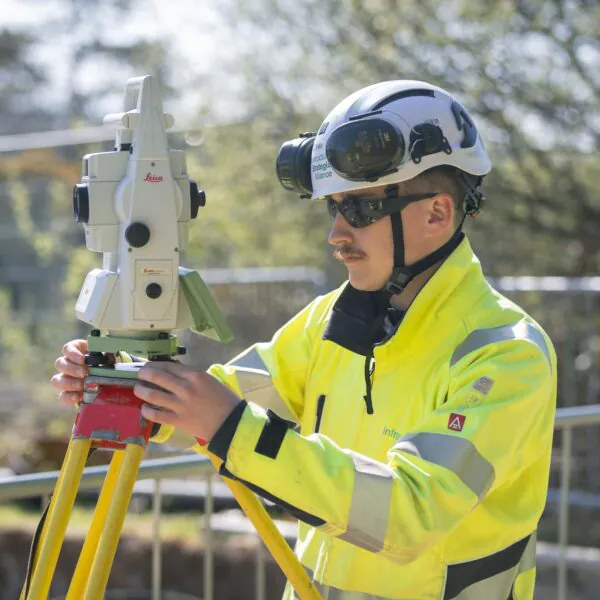 Worker in high-visibility gear uses a Leica surveying instrument mounted on a tripod, with trees and fencing in the background.