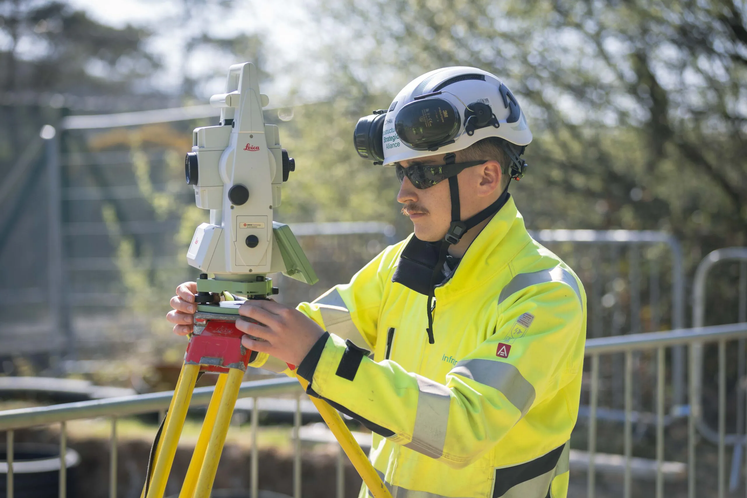 Worker in high-visibility gear uses a Leica surveying instrument mounted on a tripod, with trees and fencing in the background.