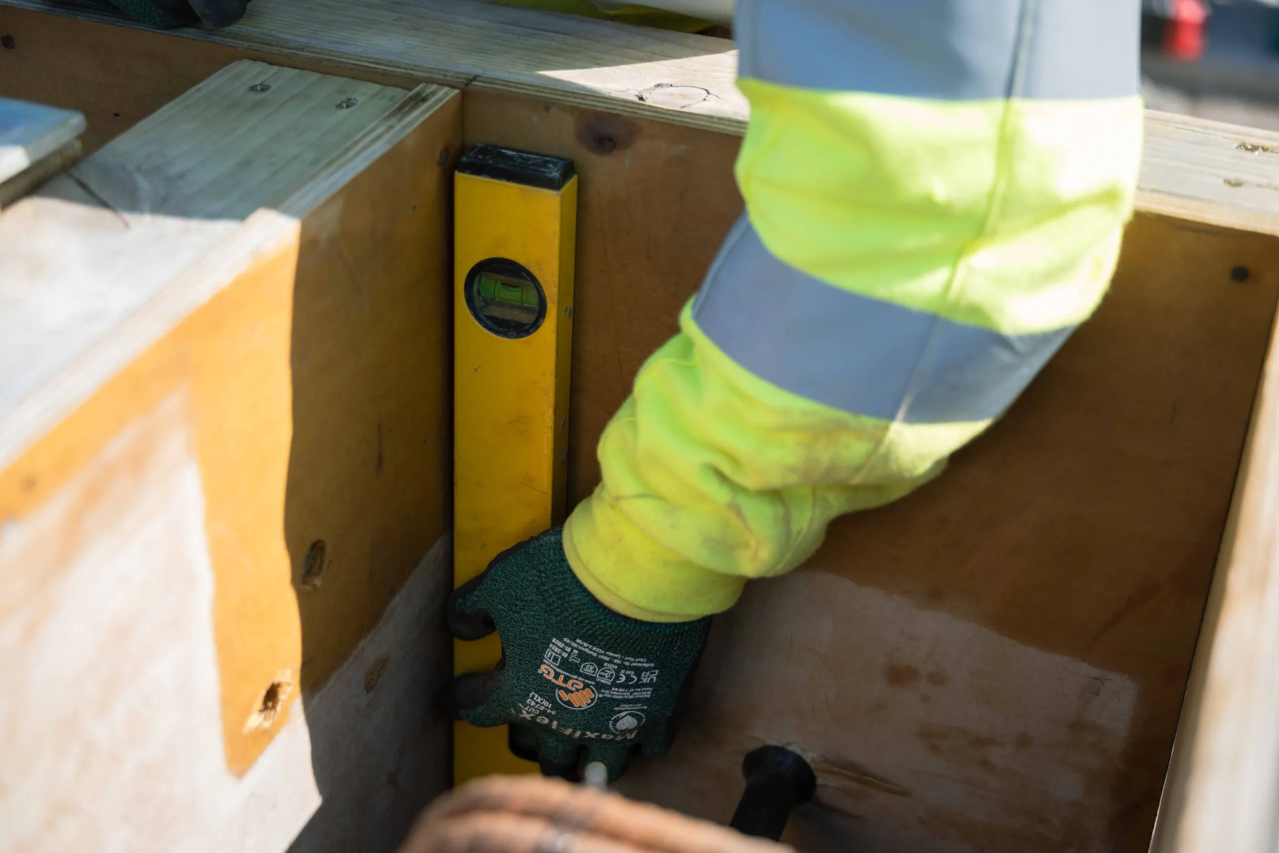 Close-up of gloved hands holding a yellow spirit level against wooden formwork at a construction site.
