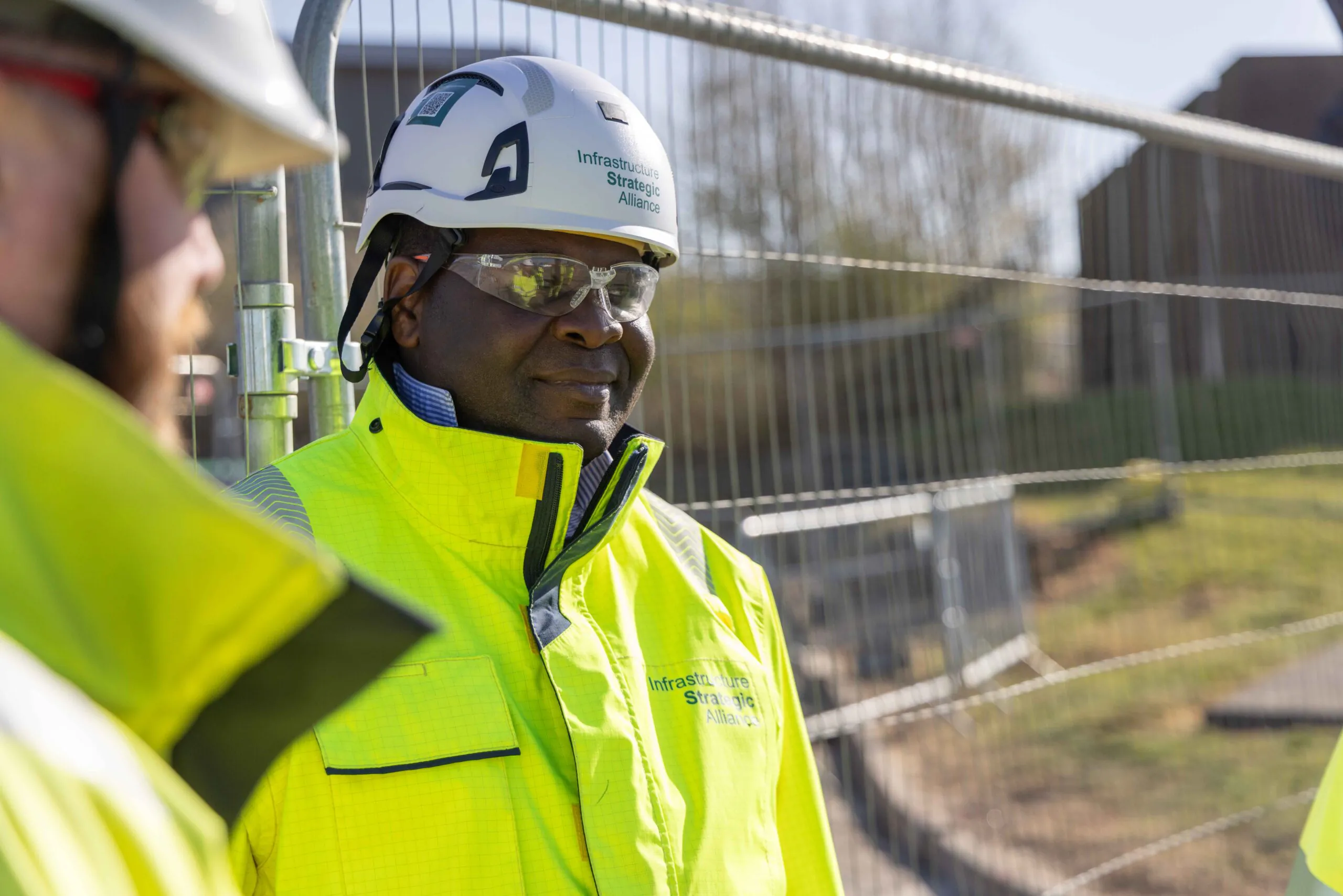Two workers in high-visibility jackets and helmets stand near fencing at an outdoor site; one faces the camera.