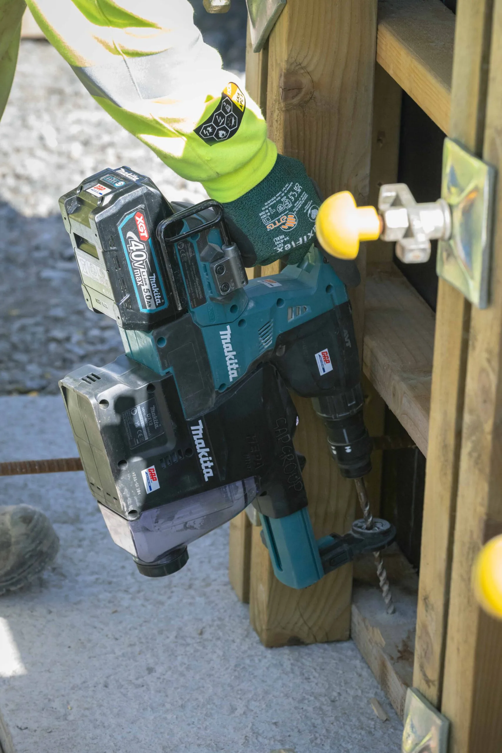 Close-up of a person using a Makita power drill to fasten components to wooden beams, wearing high-visibility safety gear and branded gloves.