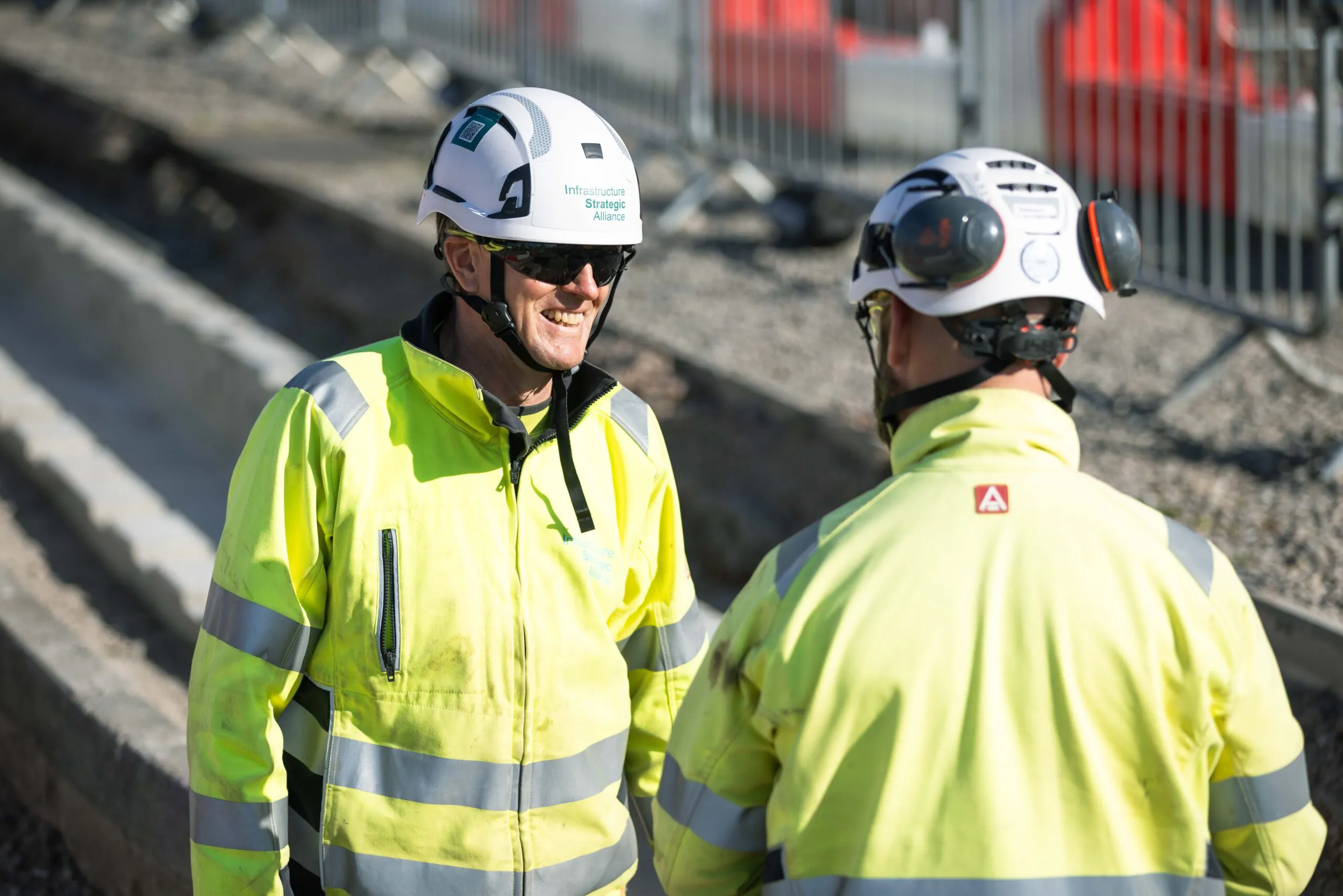 Two workers in safety gear converse at an outdoor site with safety barriers in the background.