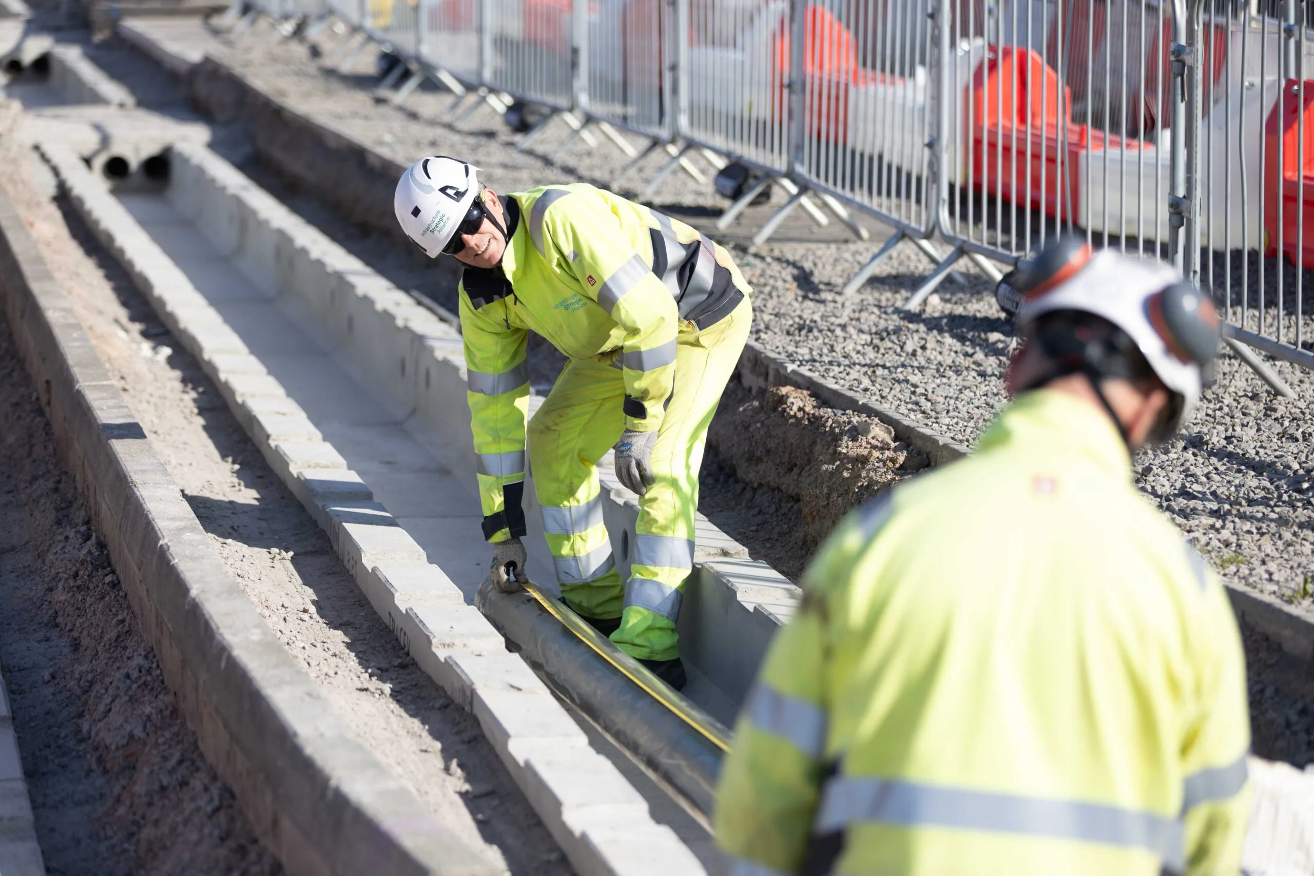Workers lay materials into trenches lined with concrete blocks, using tape measures and tools to guide placement.