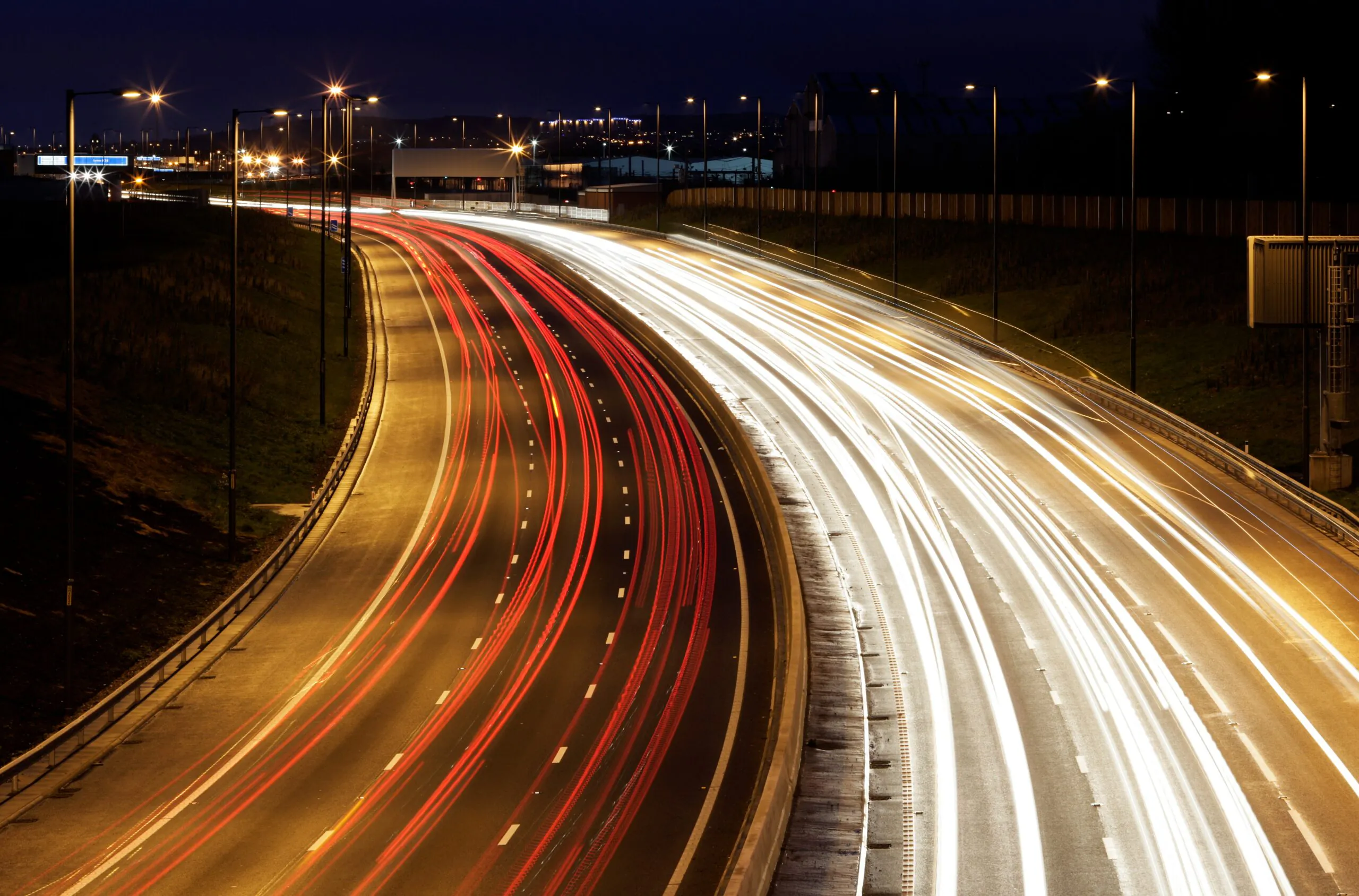 Nighttime long-exposure photo of a highway showing red and white light trails from moving vehicles, illuminated by streetlights.