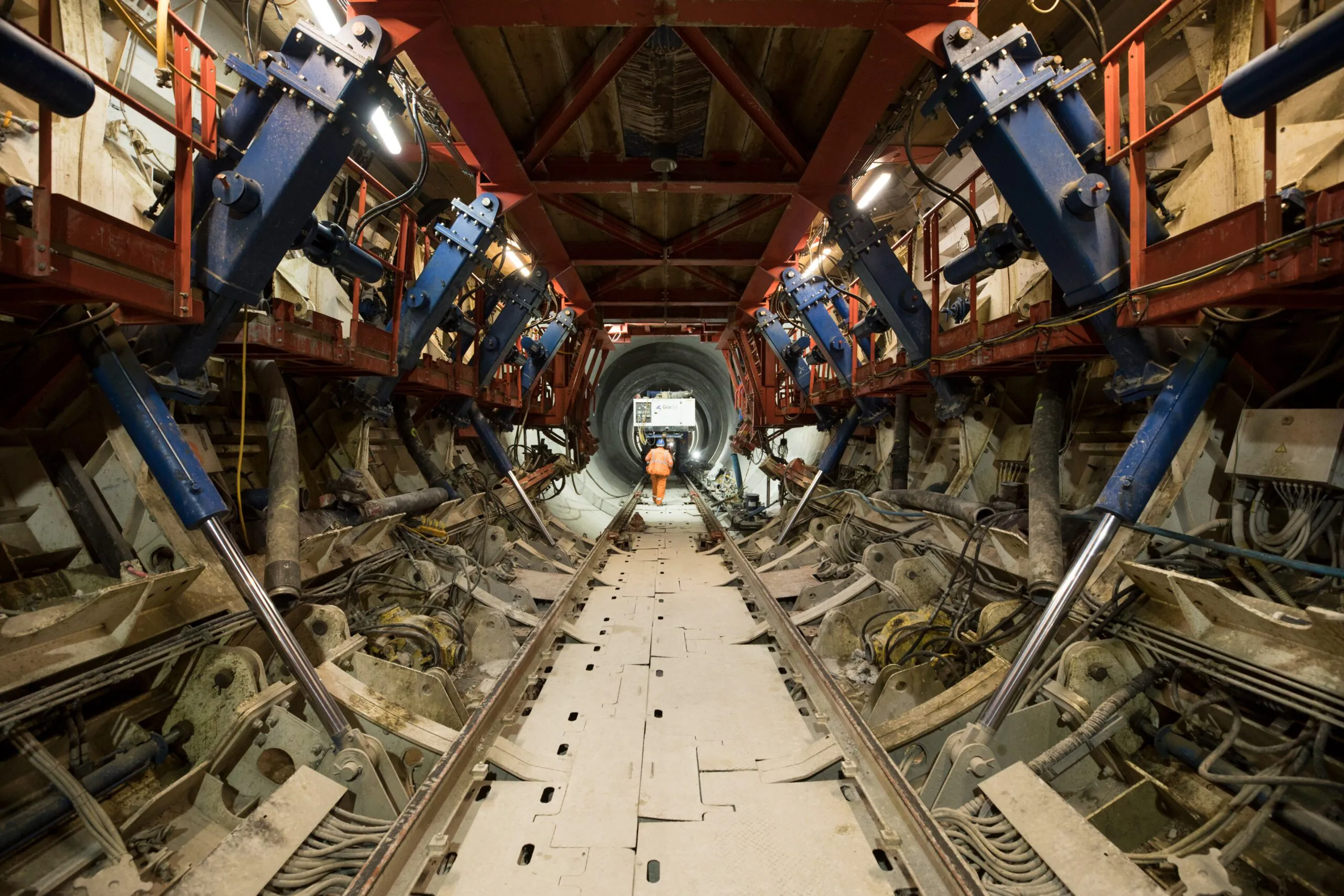 Underground tunnel under construction with complex machinery and support structures; a worker is visible at the far end.