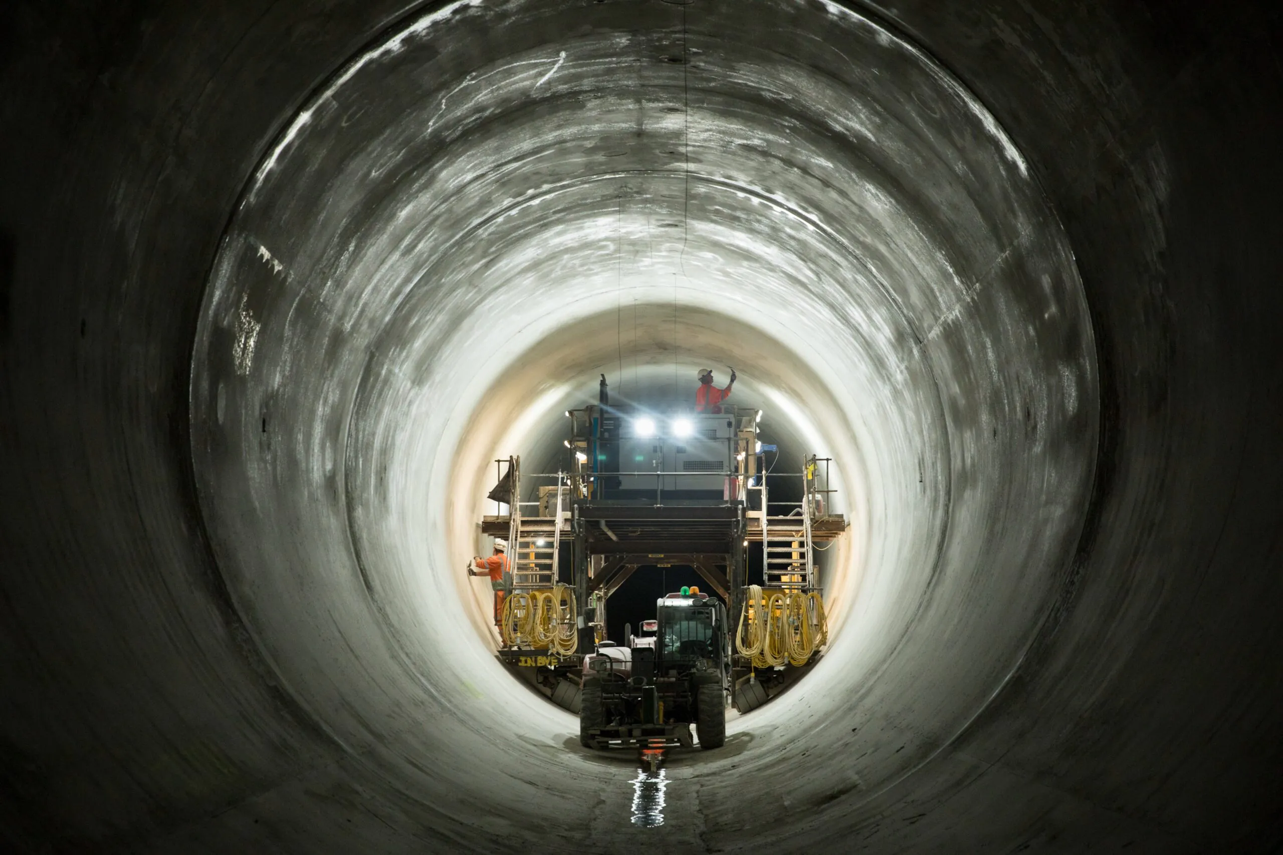 Interior of a tunnel with smooth concrete walls, brightly lit and lined with cables, pipes, and floor tracks leading deeper inside.