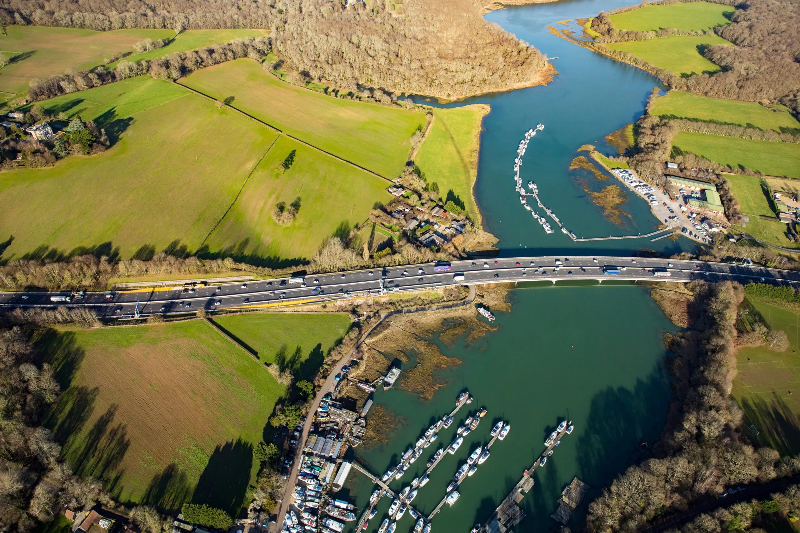 Aerial view of a bridge crossing a river, surrounded by green fields and wooded areas. Boats are docked along the riverbanks, and vehicles are visible on the bridge.