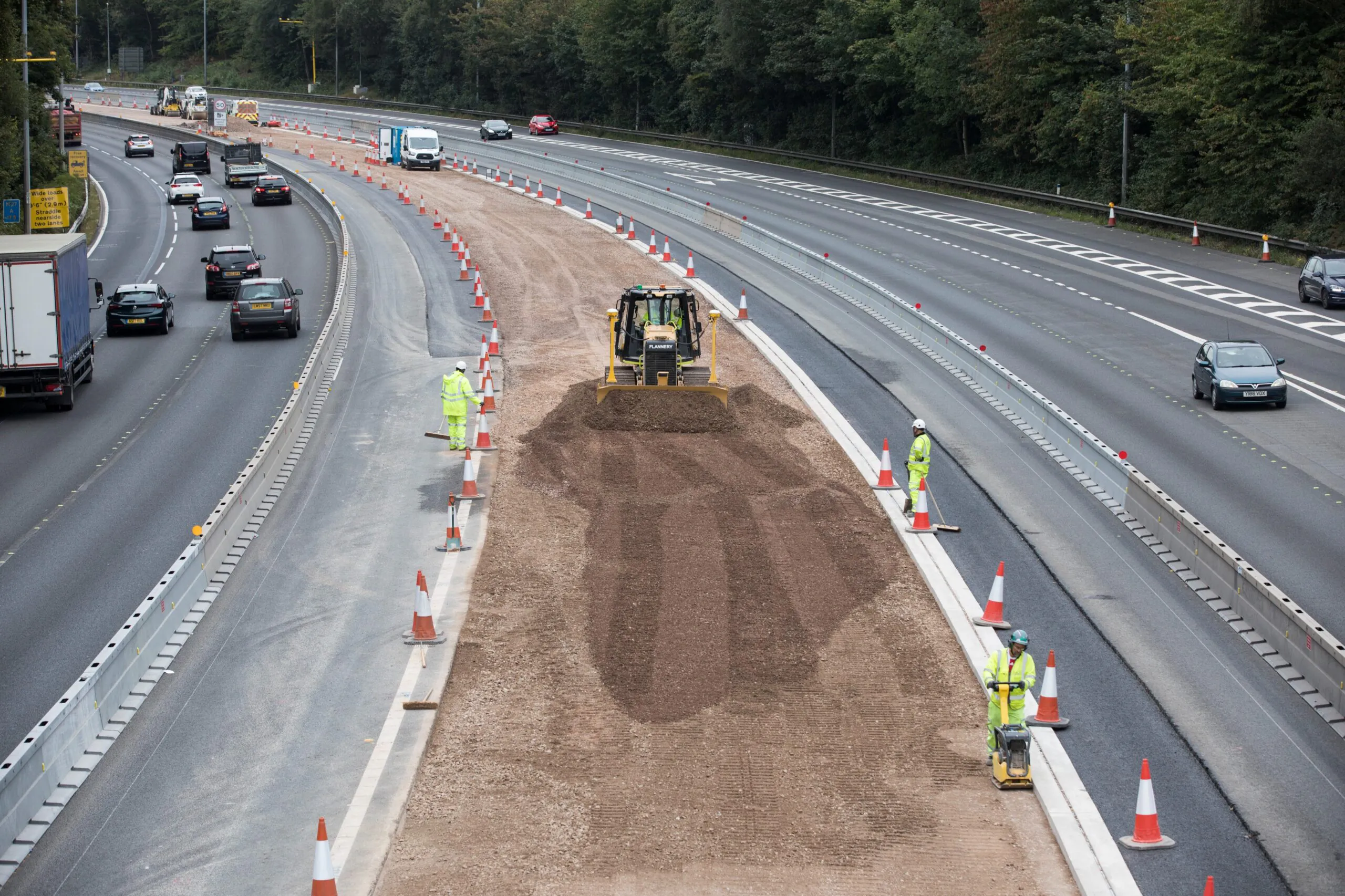 Road construction in progress with heavy machinery spreading gravel while traffic continues on adjacent lanes separated by cones and barriers.