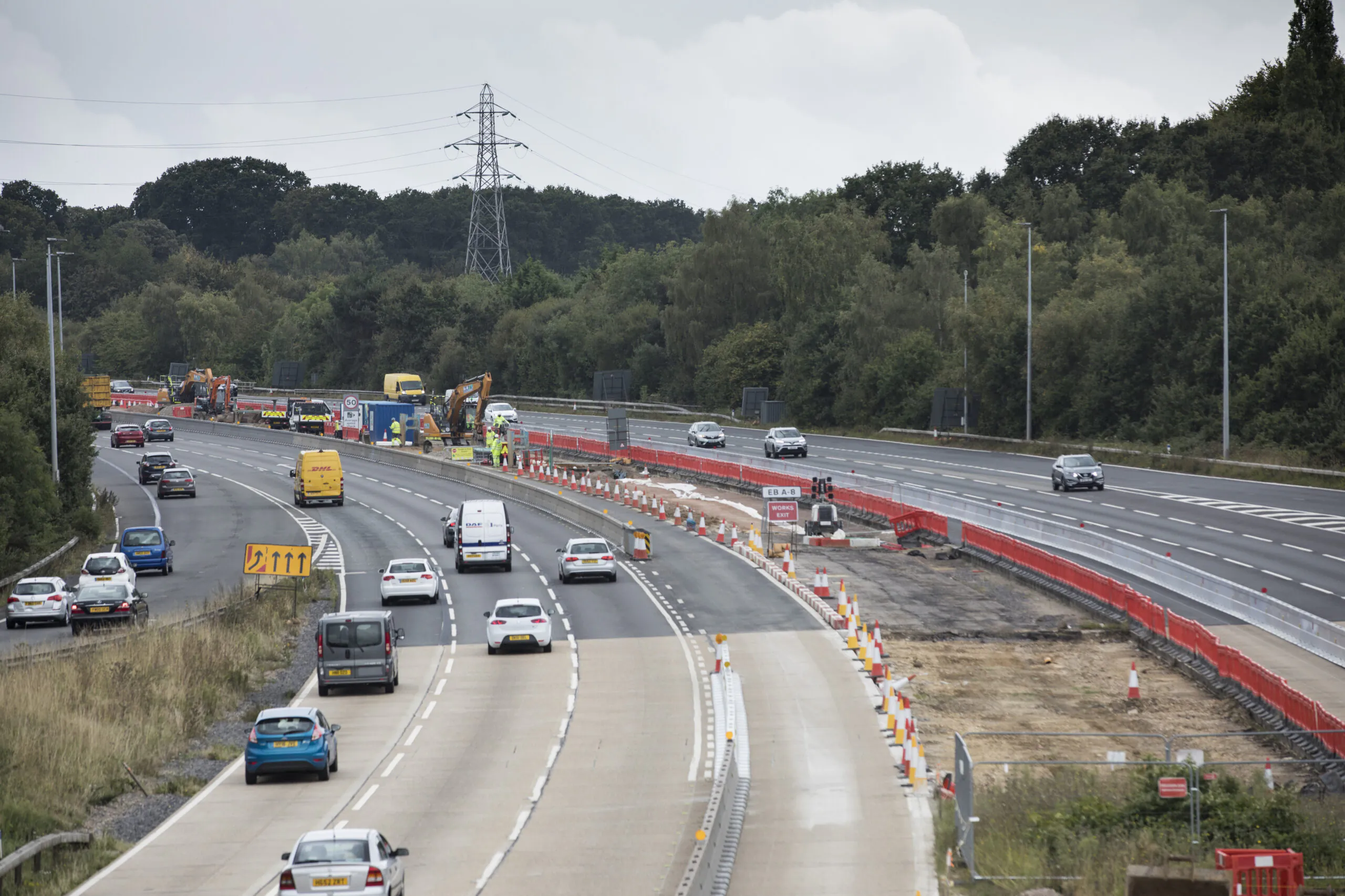 Highway expansion or repair work with traffic diverted into fewer lanes marked by orange barriers; vehicles pass through while equipment operates nearby.