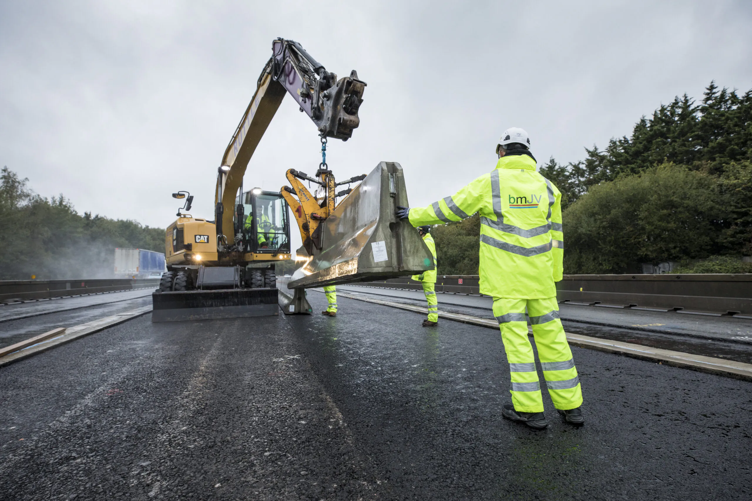 Construction workers in high-visibility clothing install large metal barriers on a freshly paved highway section using heavy machinery.