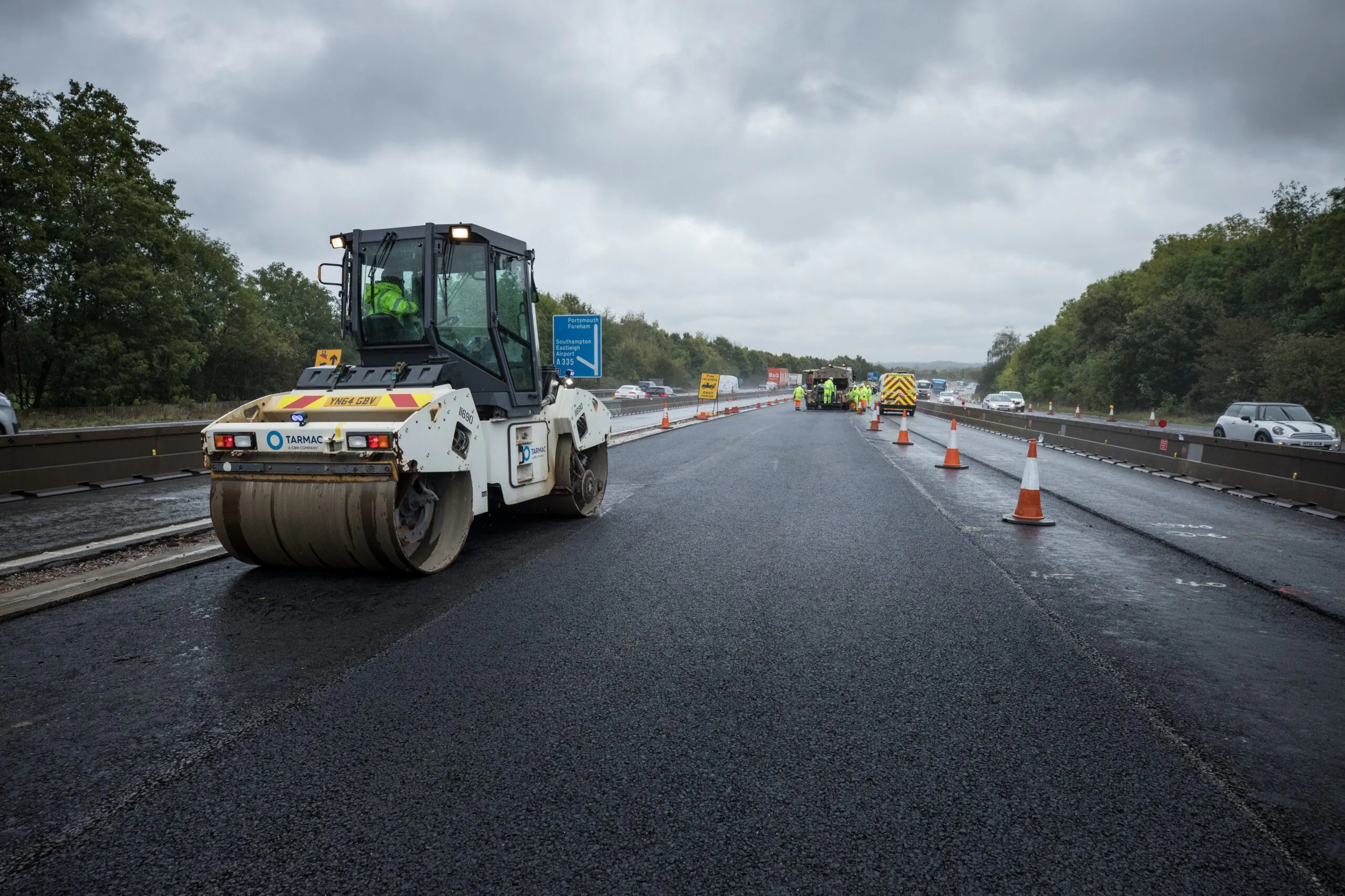 Road roller machine smoothing freshly laid asphalt on a highway under construction, with traffic cones and workers visible in the distance.