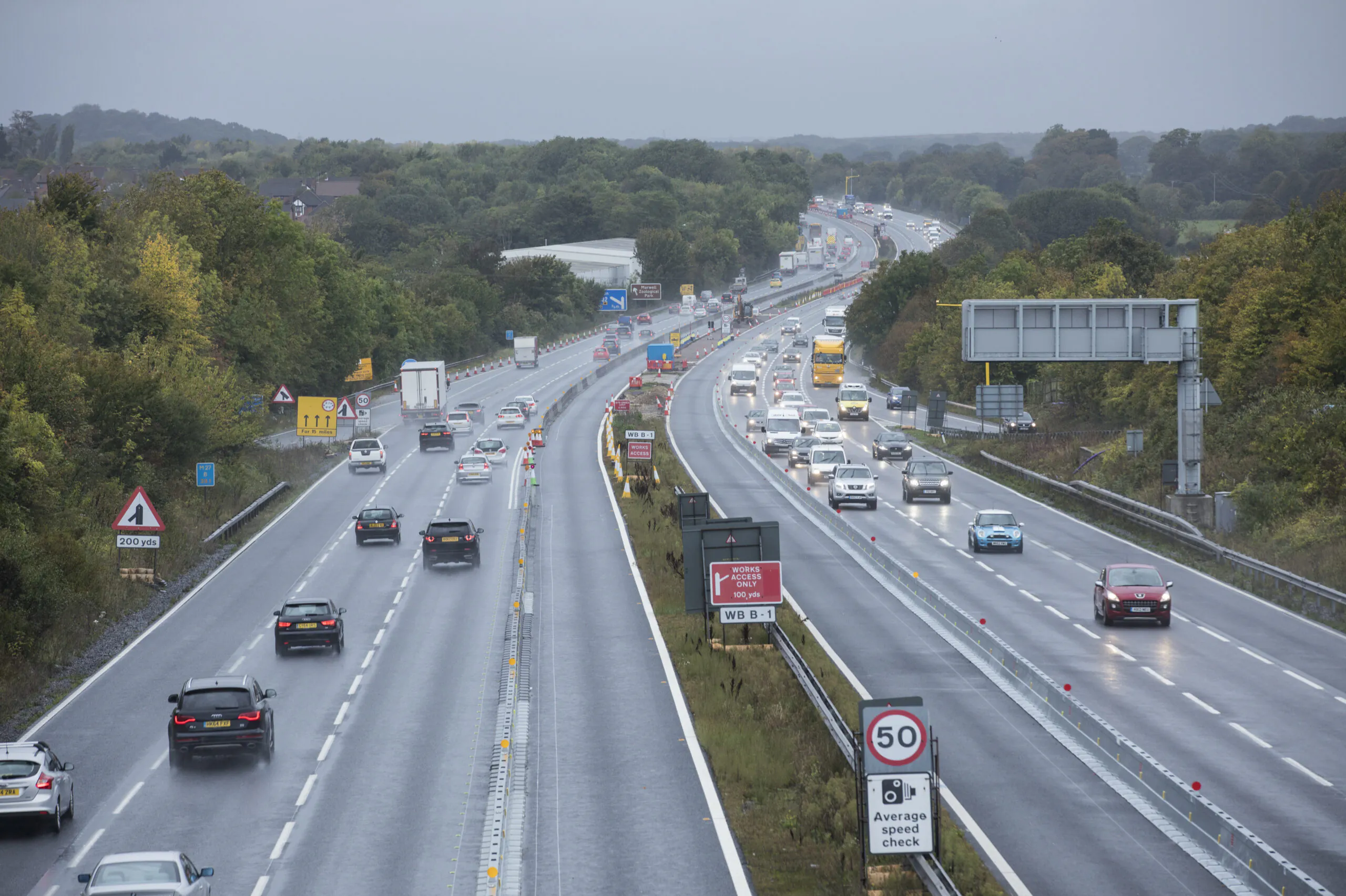 Multi-lane highway with vehicles traveling in both directions under cloudy skies; road signs indicate speed limits and lane closures due to construction.