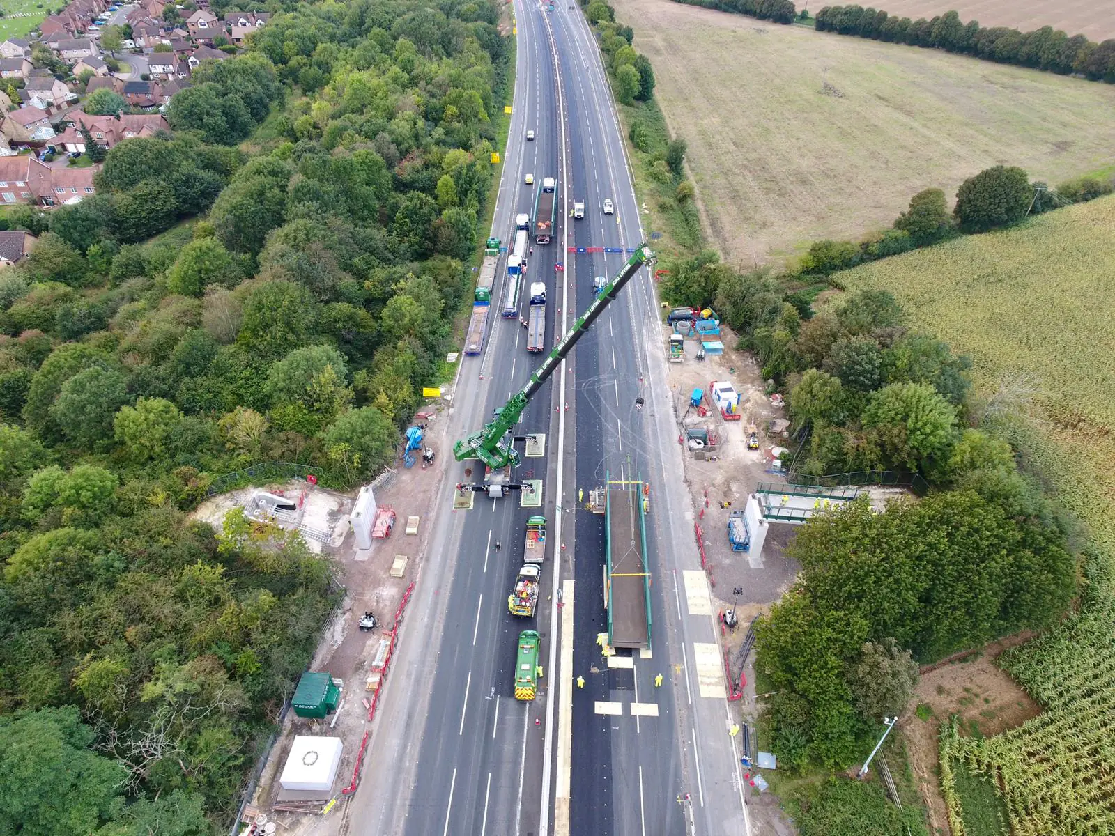 Aerial view of a highway construction site surrounded by greenery and residential areas, with heavy machinery including a crane in operation.