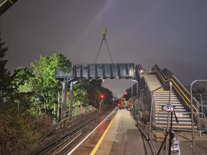 Nighttime railway construction scene with workers using cranes to install or remove footbridge sections at an active train station.