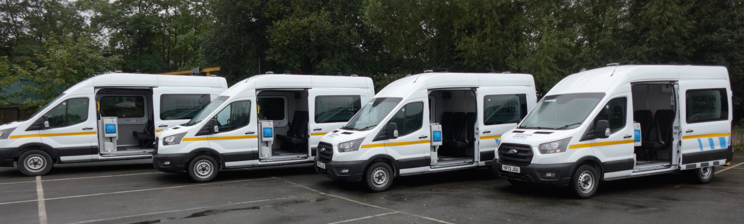 Four white welfare vans branded "Morgan Sindall" with open doors, lined up in a parking lot surrounded by trees.