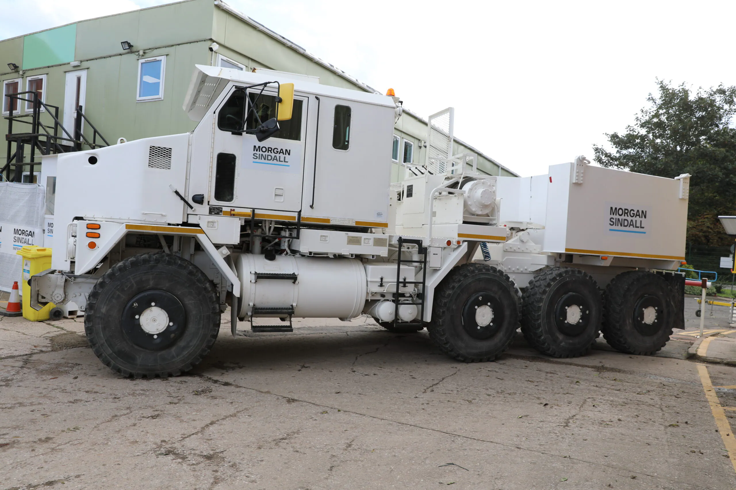 Large off-road vehicle marked "Morgan Sindall" with six wheels and heavy-duty rear equipment, parked outside industrial buildings.