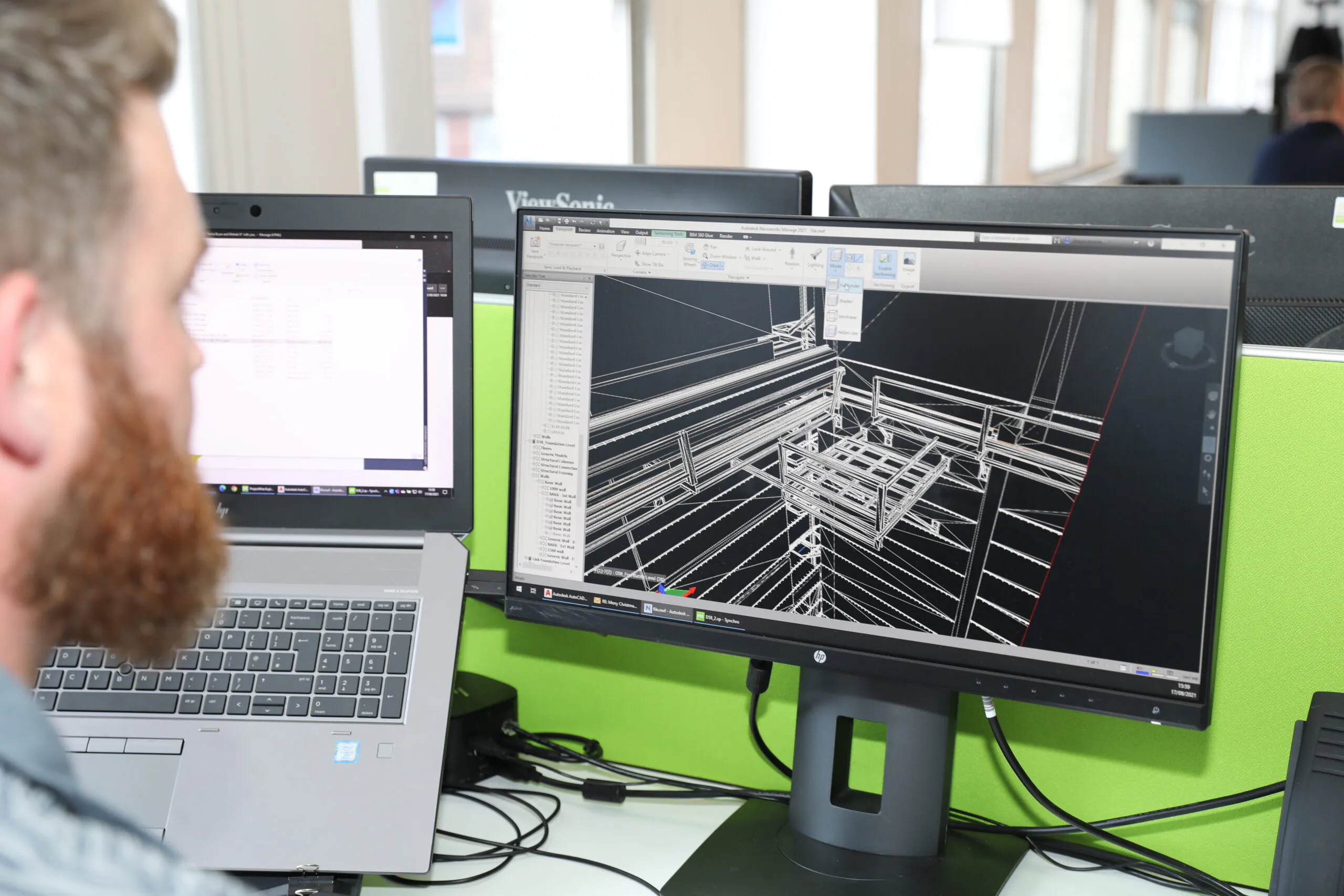 Office setting with a person working at a computer displaying 3D architectural designs, with a laptop open beside it.