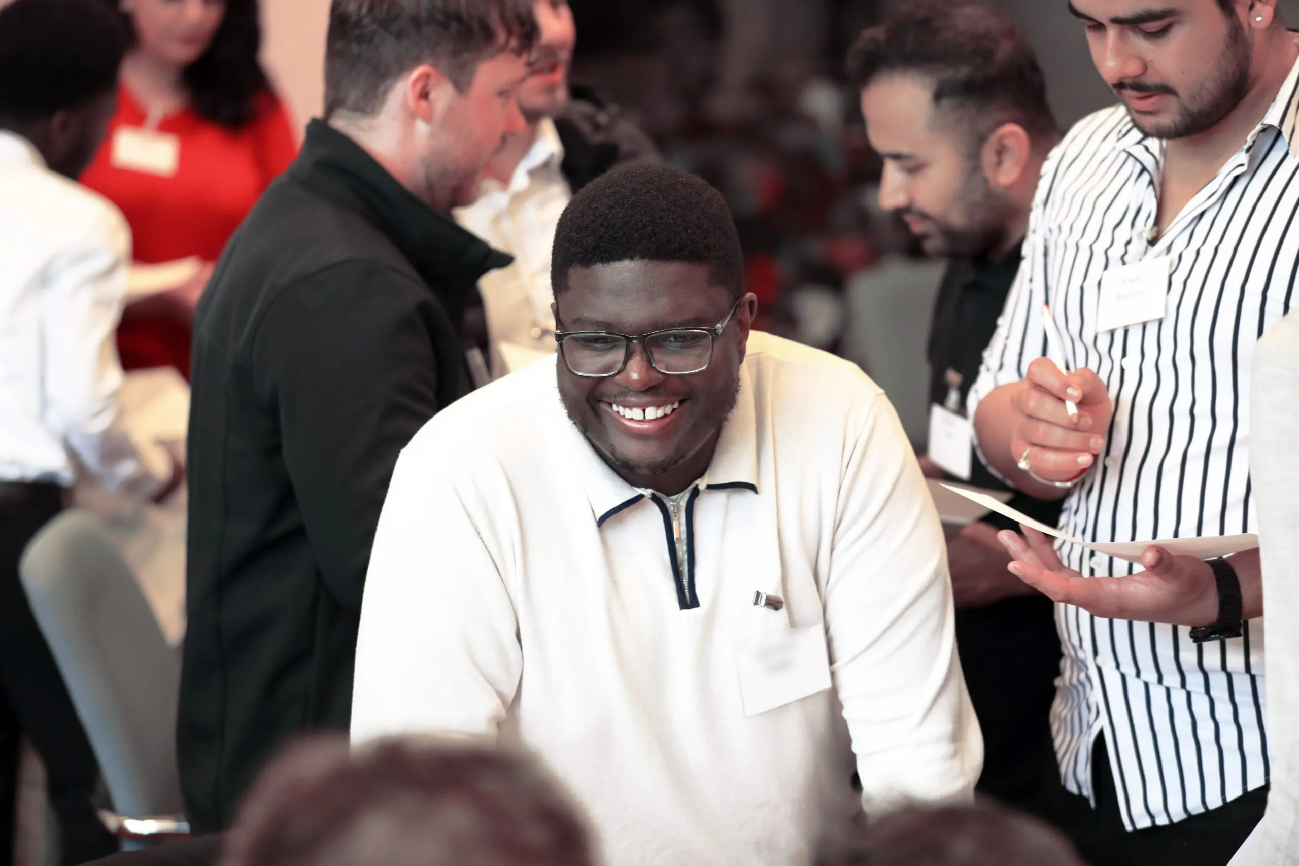 Group of people gathered indoors at a social or professional event; one person wearing glasses and a light shirt is smiling while conversing.