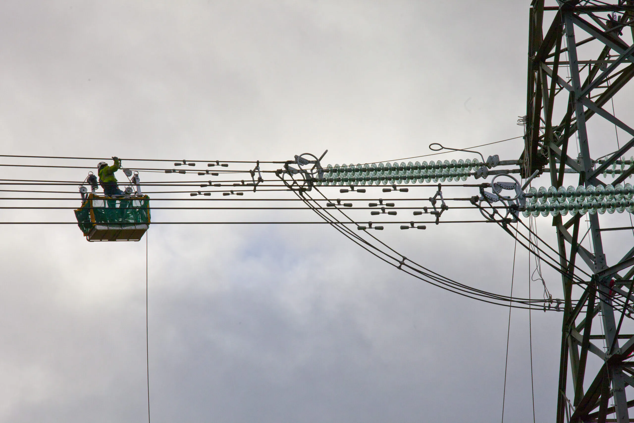 Workers in safety gear suspended on power lines using a green basket lift attached to an electrical tower, performing maintenance against a cloudy sky.