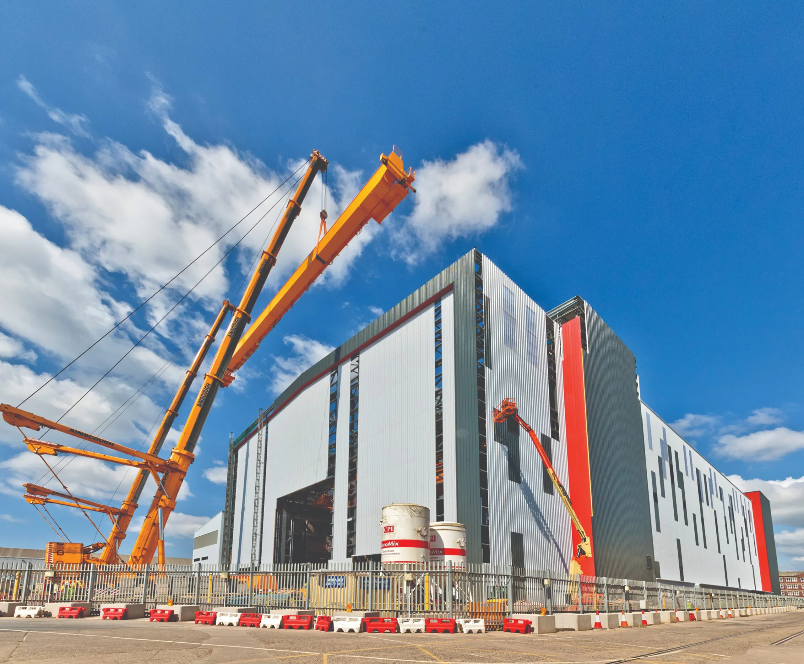 Large industrial building under construction or maintenance with multiple yellow cranes operating around it under a bright blue sky.
