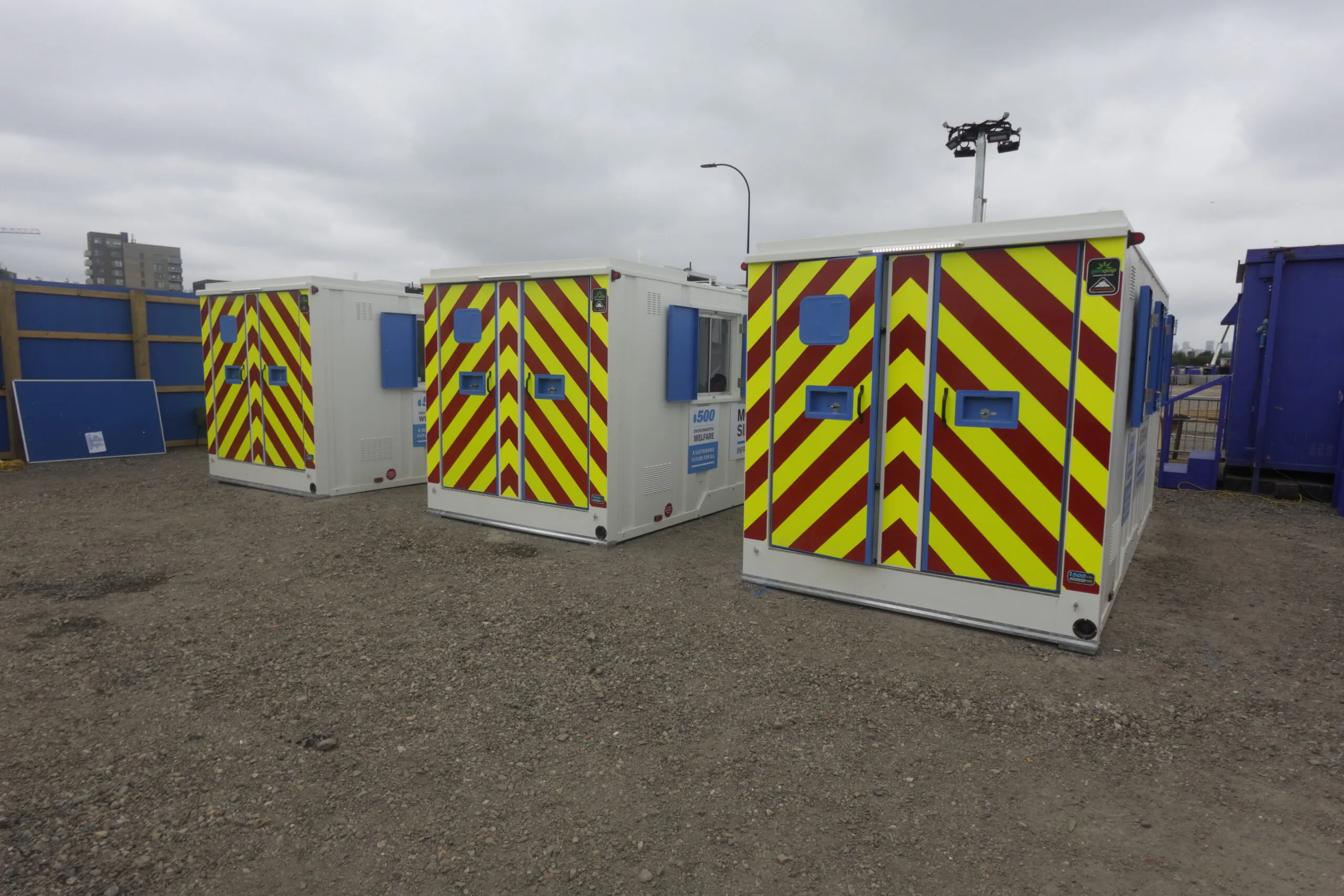Three portable cabins with white exteriors and red-yellow chevron patterns on their doors, parked on gravel under an overcast sky.