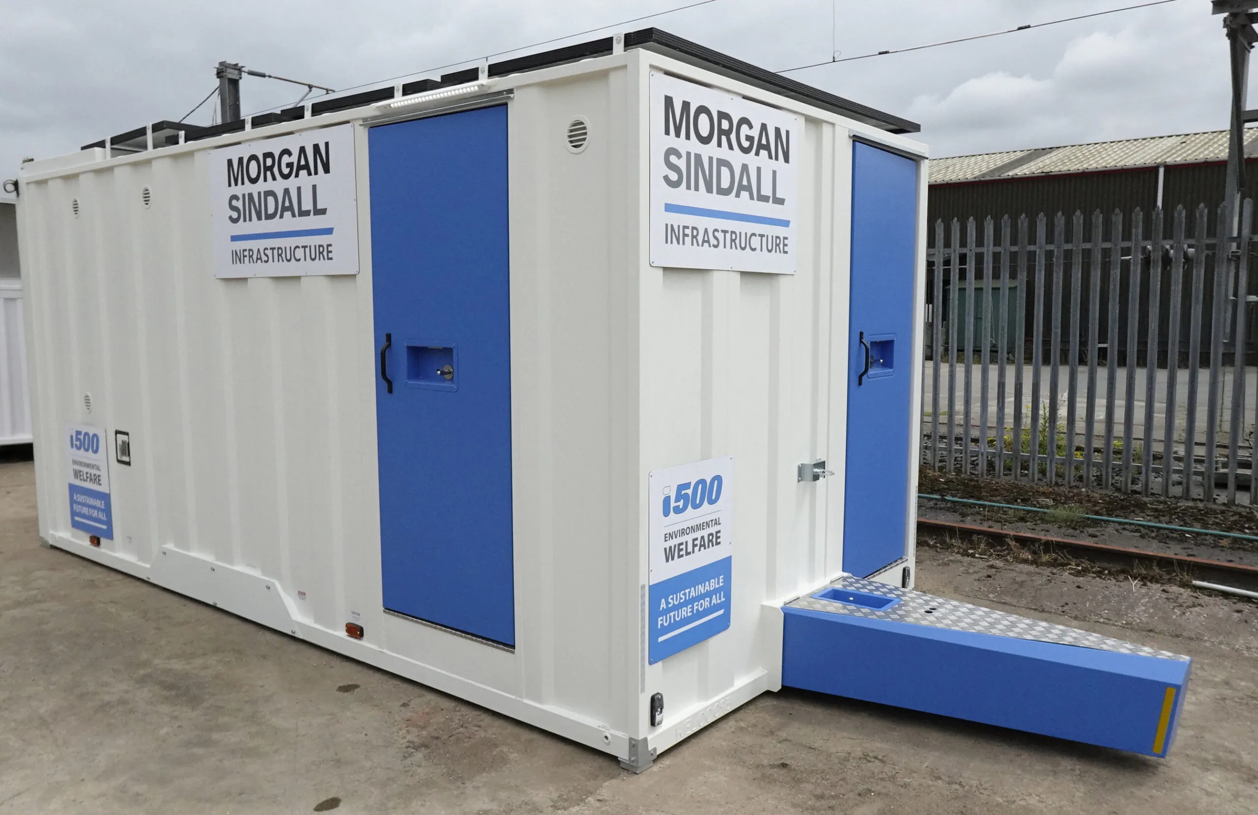 Standalone white welfare unit with blue doors and windows, placed on gravel at a construction site, featuring signage for the i500 environmental welfare initiative.
