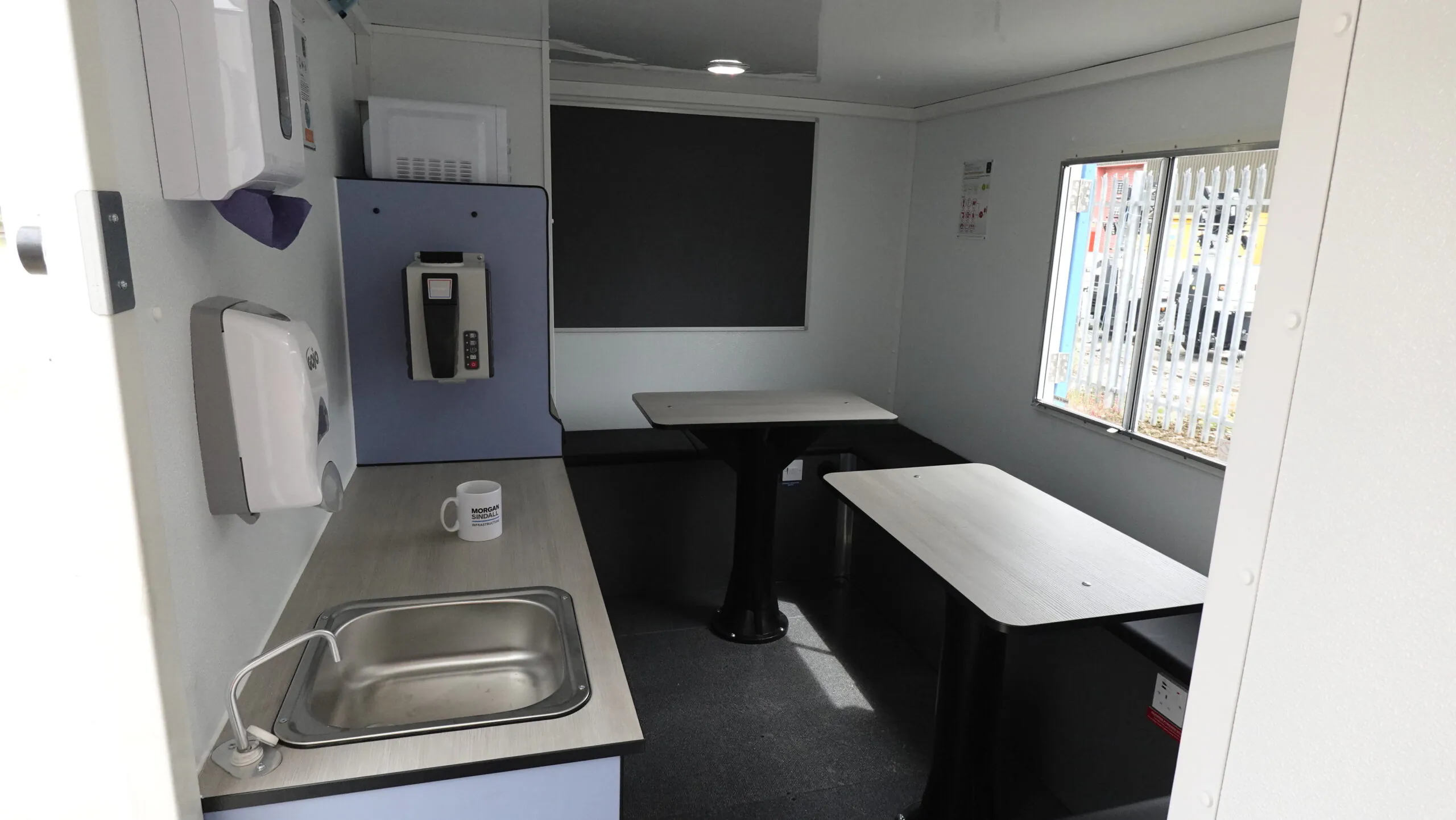Interior of a compact welfare unit featuring a kitchenette with sink, countertop, wall-mounted dispensers, and seating area with tables and benches. A window provides natural light.