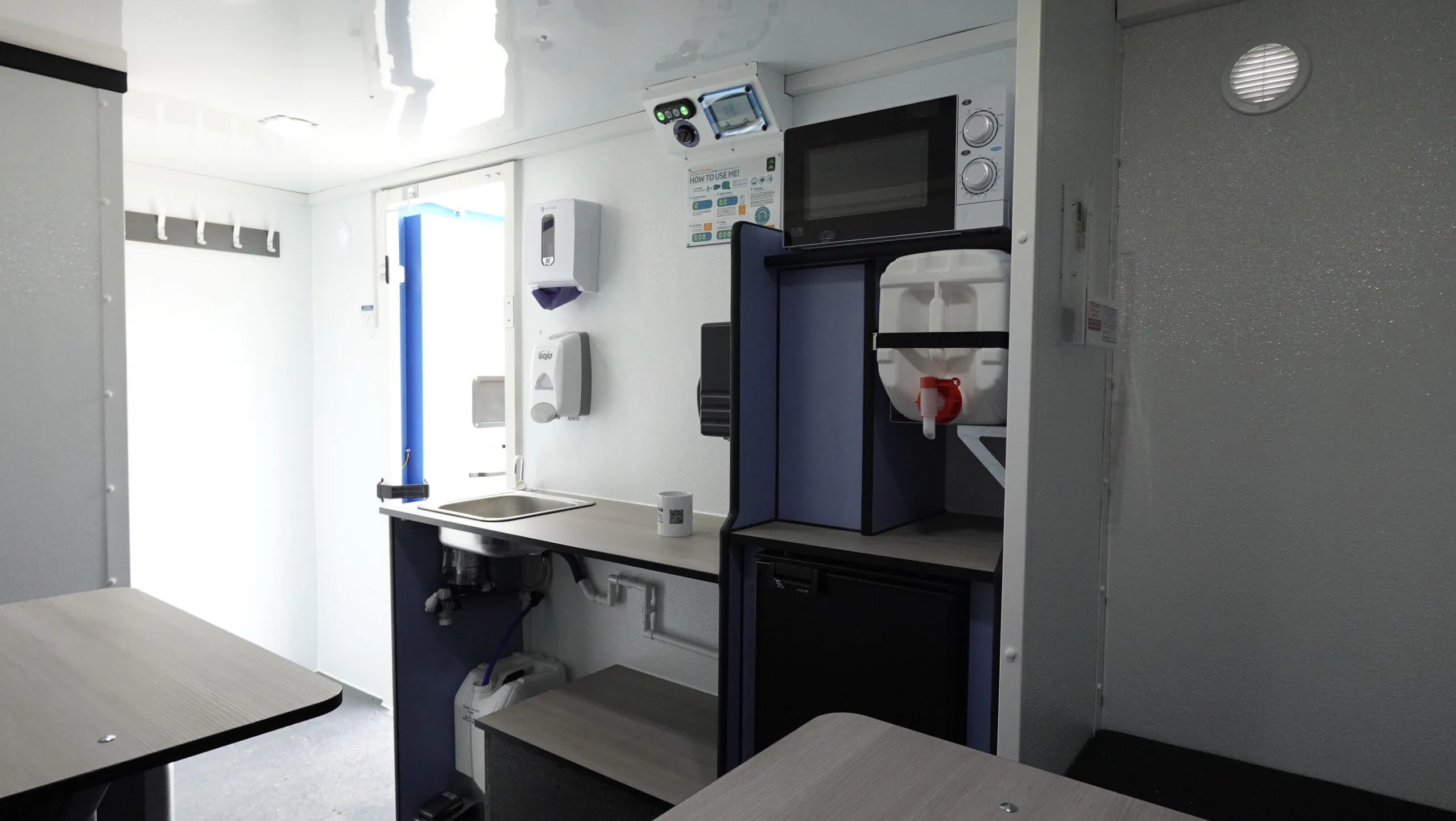 Alternate view of the welfare unit interior showing a microwave above the counter, wall-mounted dispensers, and a door leading outside.