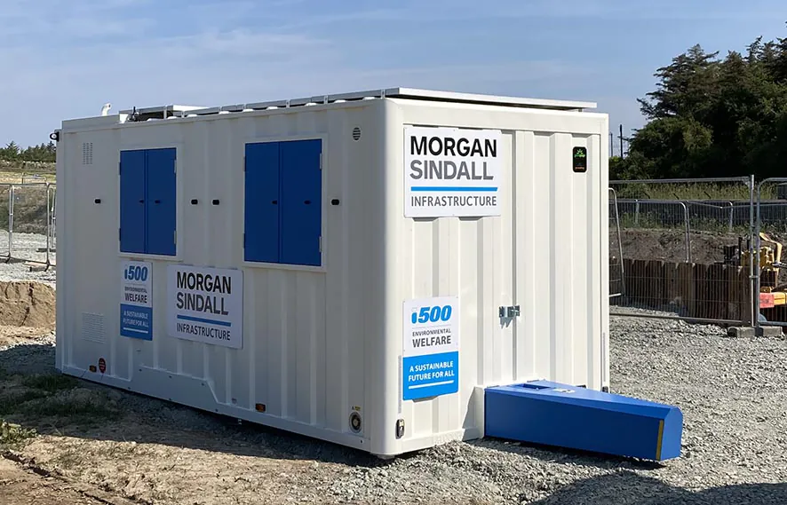 Standalone white welfare unit with blue doors and windows, placed on gravel at a construction site, featuring signage for the i500 environmental welfare initiative.