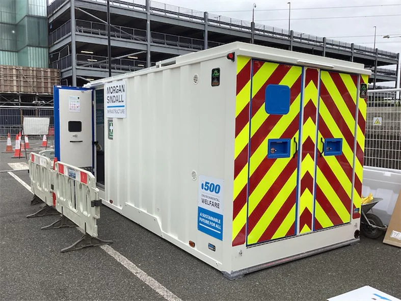 Portable cabin labeled “MORGAN SINDALL INFRASTRUCTURE” with red-yellow chevron doors, parked near construction barriers in an urban area.