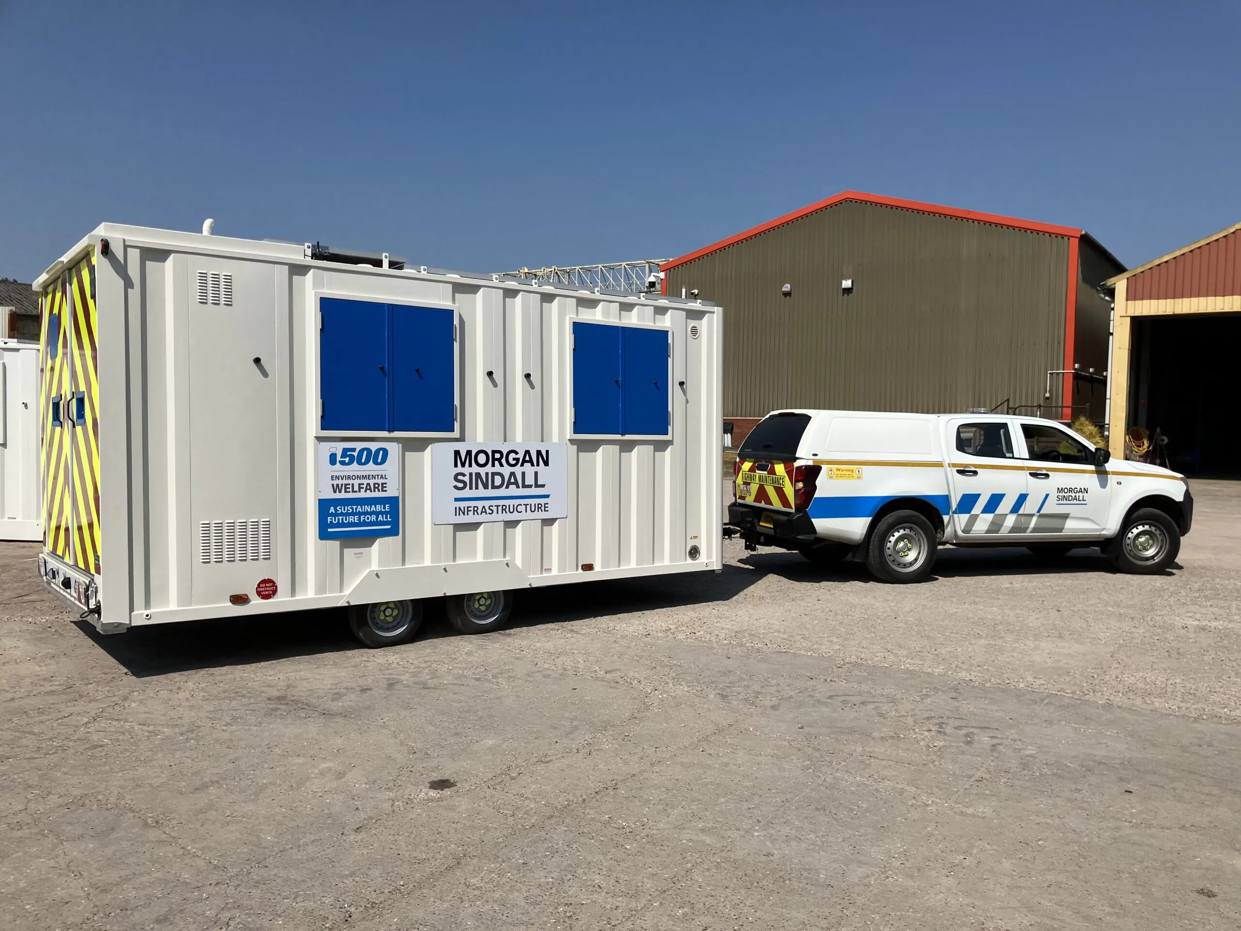 White mobile welfare unit with blue doors and windows labeled "Morgan Sindall Infrastructure," being towed by a utility vehicle in an industrial area.