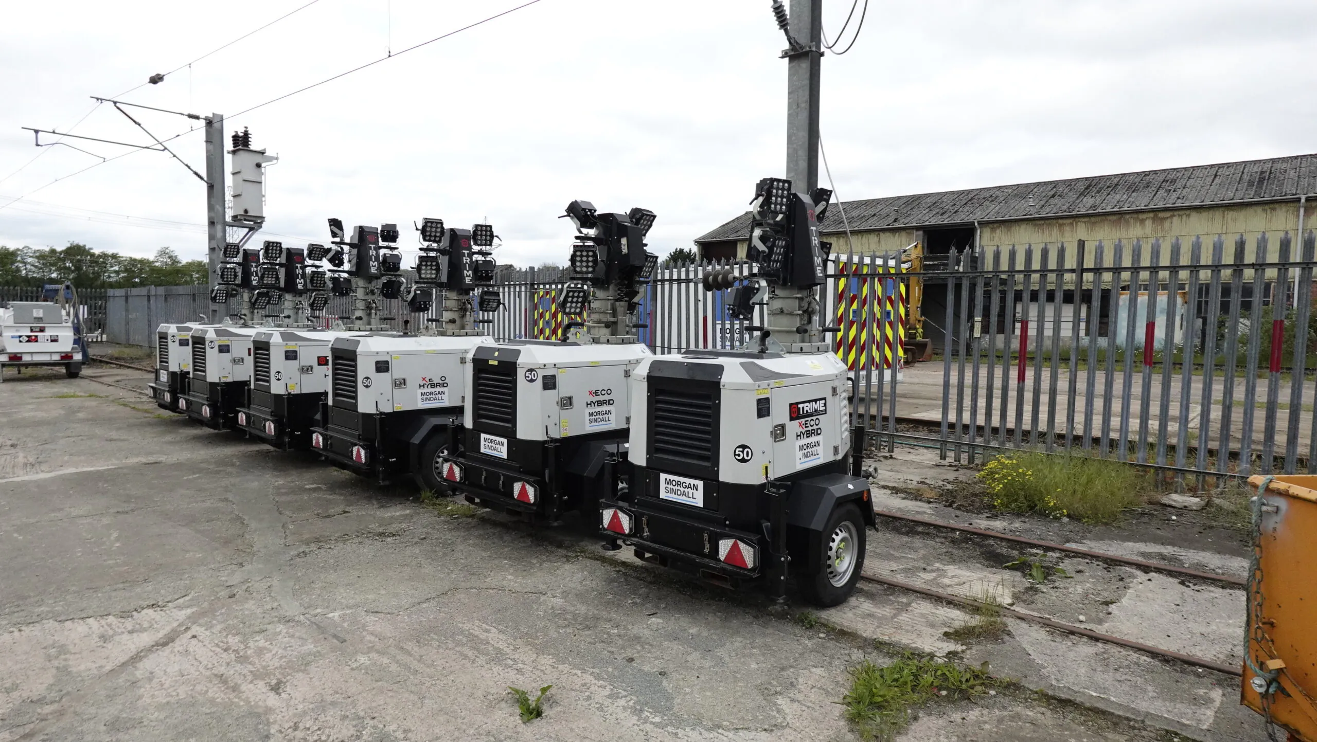 Multiple portable tower lights labeled "Trime" and "Morgan Sindall" lined up outdoors near industrial buildings.