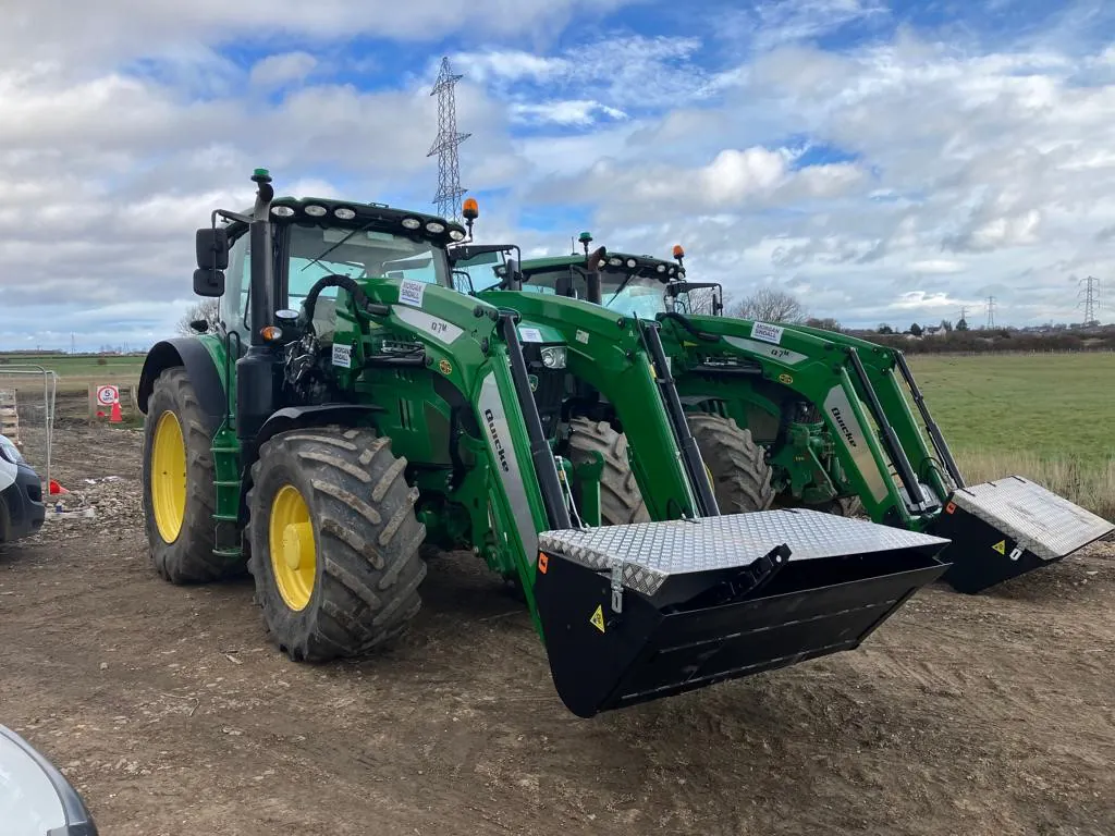 Two green John Deere tractors with front loaders and large yellow wheels parked side by side on a dirt path in an open field.