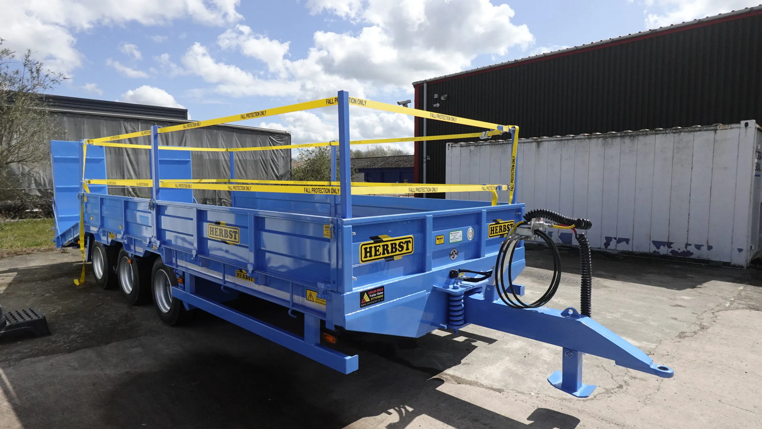 Blue low-loader trailer labeled "Herbst" with safety railings, parked outdoors under a partly cloudy sky.