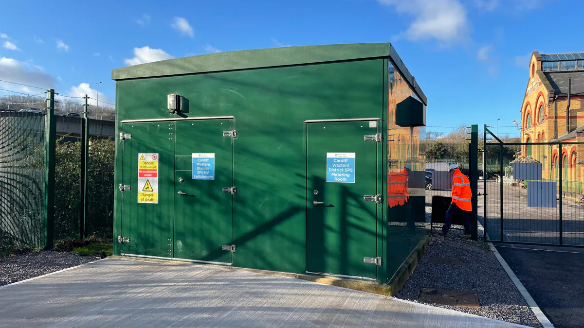 Green utility building with two doors and warning signs, surrounded by fencing. A person in high-visibility clothing walks nearby under a clear blue sky.