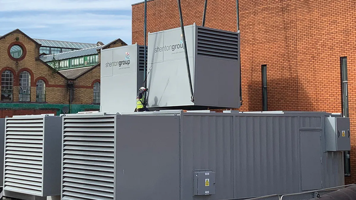 Installation of grey generator units labeled "Shenton Group" using cranes outside a brick building.