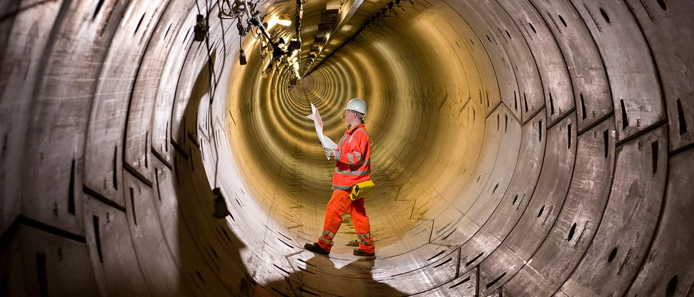 Person in high-visibility orange gear and hard hat standing inside a large, well-lit concrete tunnel, holding construction plans.