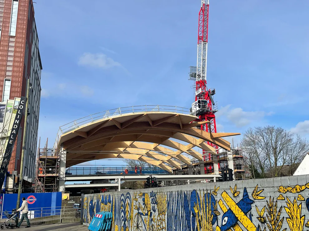 Daytime view of a construction site with a large wooden curved roof, scaffolding, cranes, and colorful murals on nearby walls.
