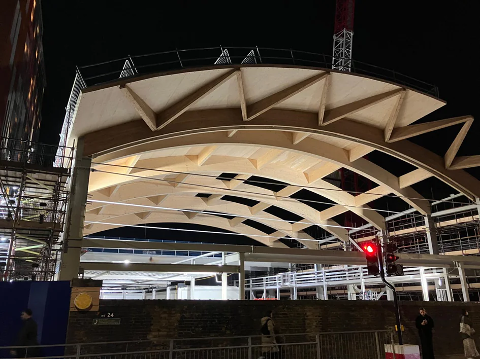 Nighttime image of a building under construction with an illuminated curved roof supported by beams and surrounded by scaffolding.
