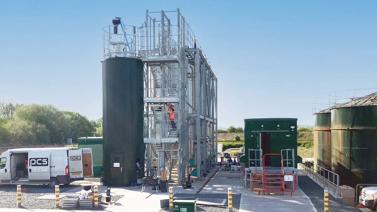 Industrial site with tall metal structures and tanks connected by pipes; workers and green utility buildings visible under clear skies.