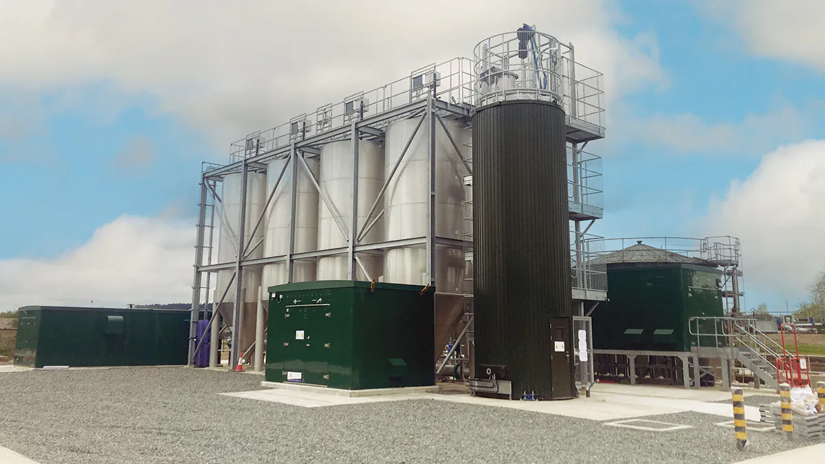 Industrial site showing construction activity with workers, machinery, and green utility buildings under a partly cloudy sky.